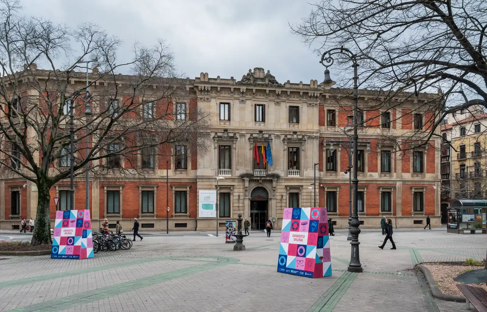 Fachada del Parlamento de Navarra en Pamplona, un palacio de ladrillo rojo y piedra con las banderas de Navarra, España y la Unión Europea sobre la puerta principal, vista desde la plaza en un día nublado de invierno.
