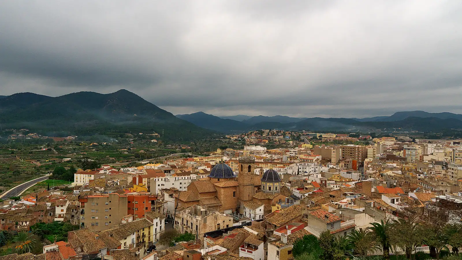 Vista aérea del casco urbano de Onda, con la iglesia de tejados de cerámica azul en el centro y bloques de viviendas alrededor, al pie de una sierra de laderas boscosas bajo un cielo encapotado.