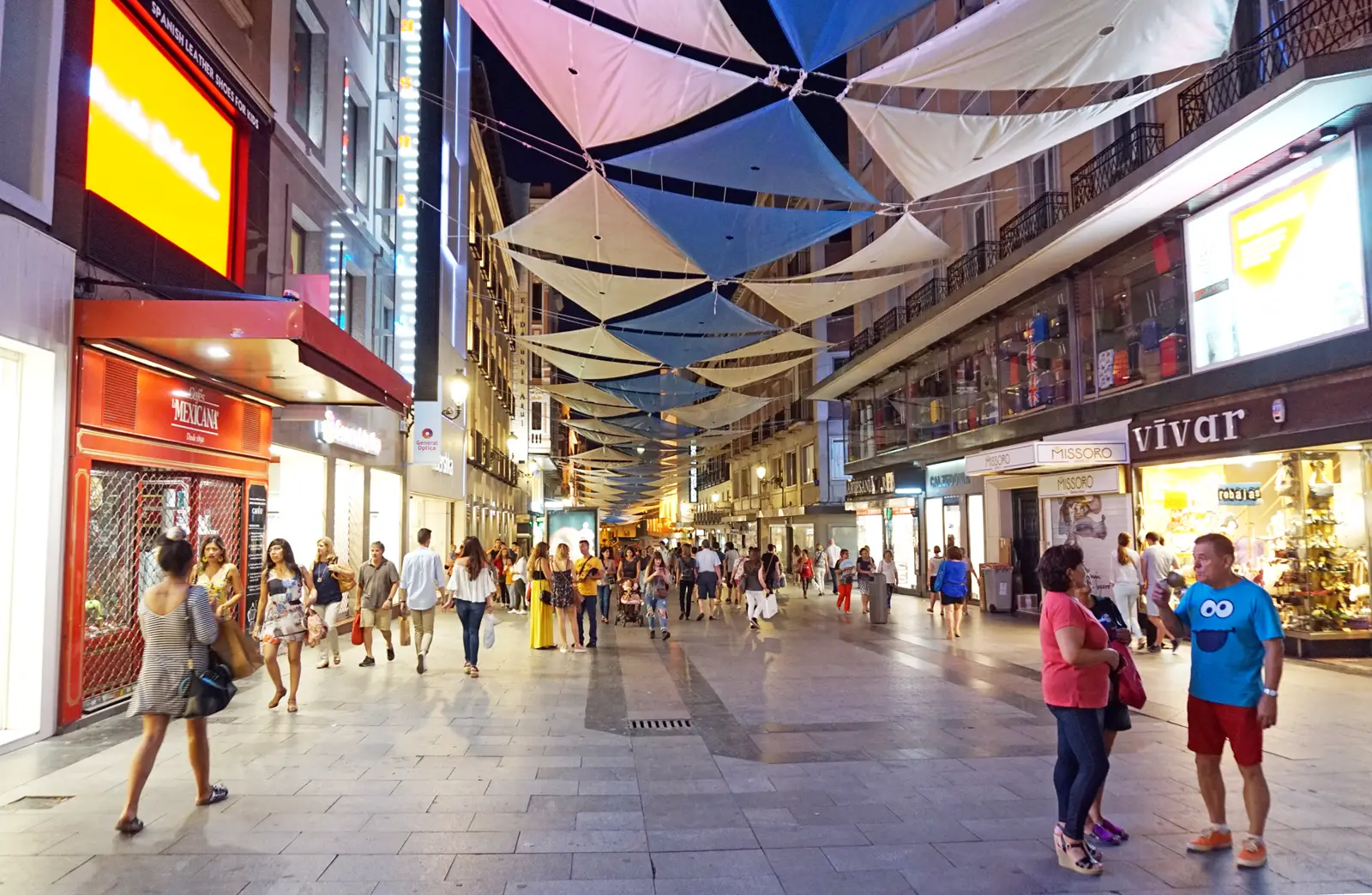 Calle peatonal de Madrid al anochecer, con escaparates iluminados a ambos lados, toldos triangulares colgados sobre la vía y decenas de personas caminando con bolsas de compra