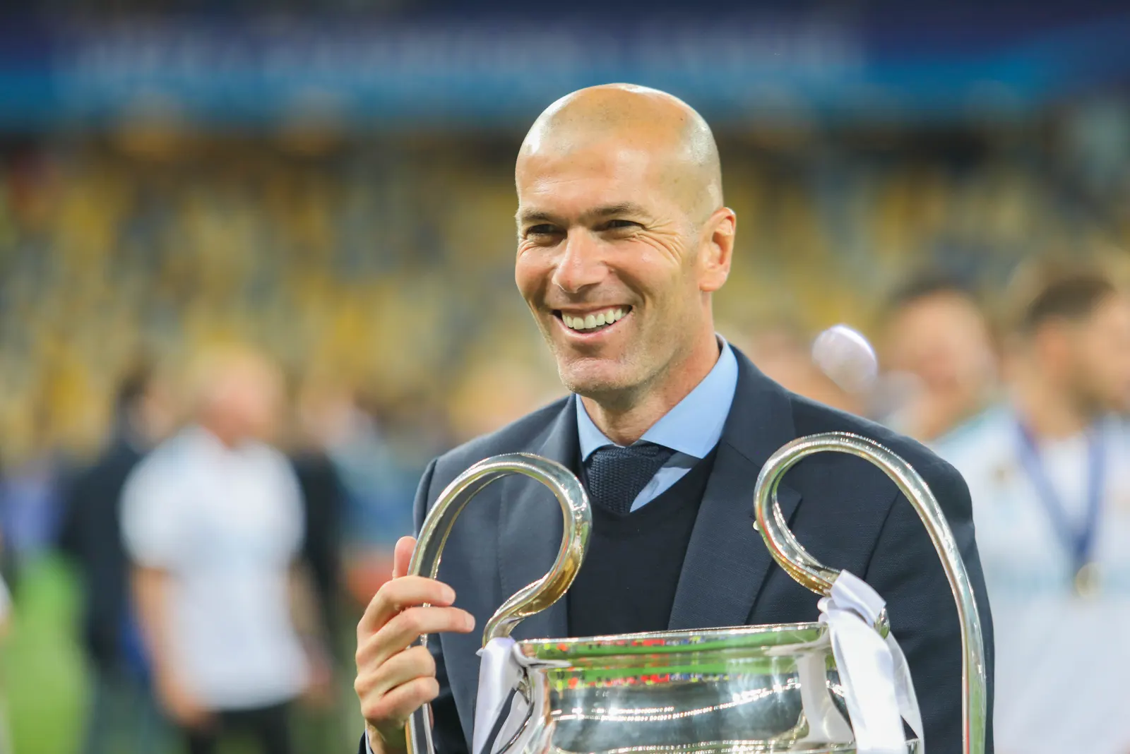 Zinedine Zidane, de traje oscuro y camisa azul, sonríe sobre el césped con las dos manos en las asas del trofeo de la Liga de Campeones