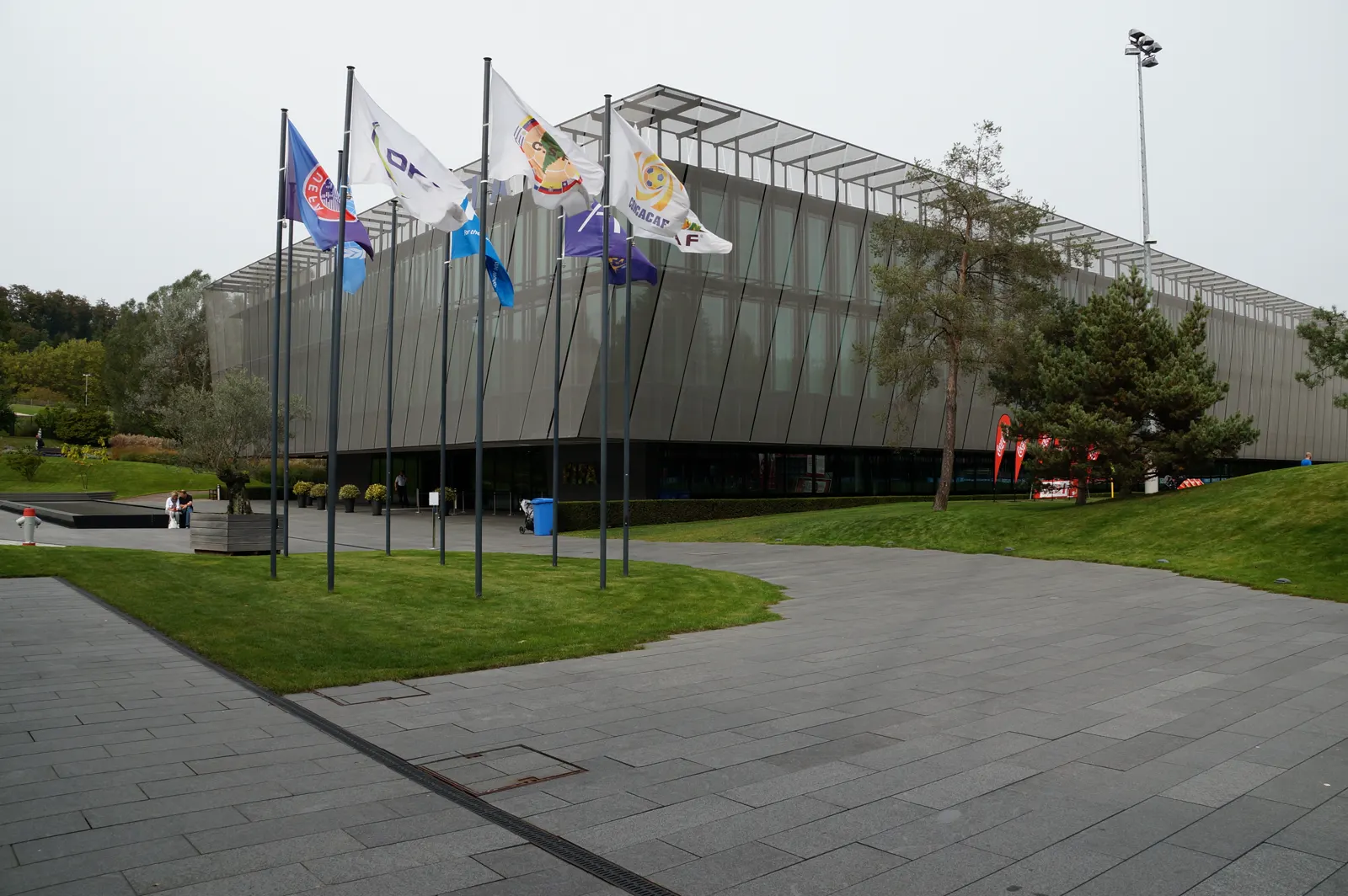 Edificio acristalado y de perfil inclinado de la sede de la FIFA en Zúrich, rodeado de césped y con siete mástiles en primer plano con banderas, entre ellas las de la UEFA y la Concacaf