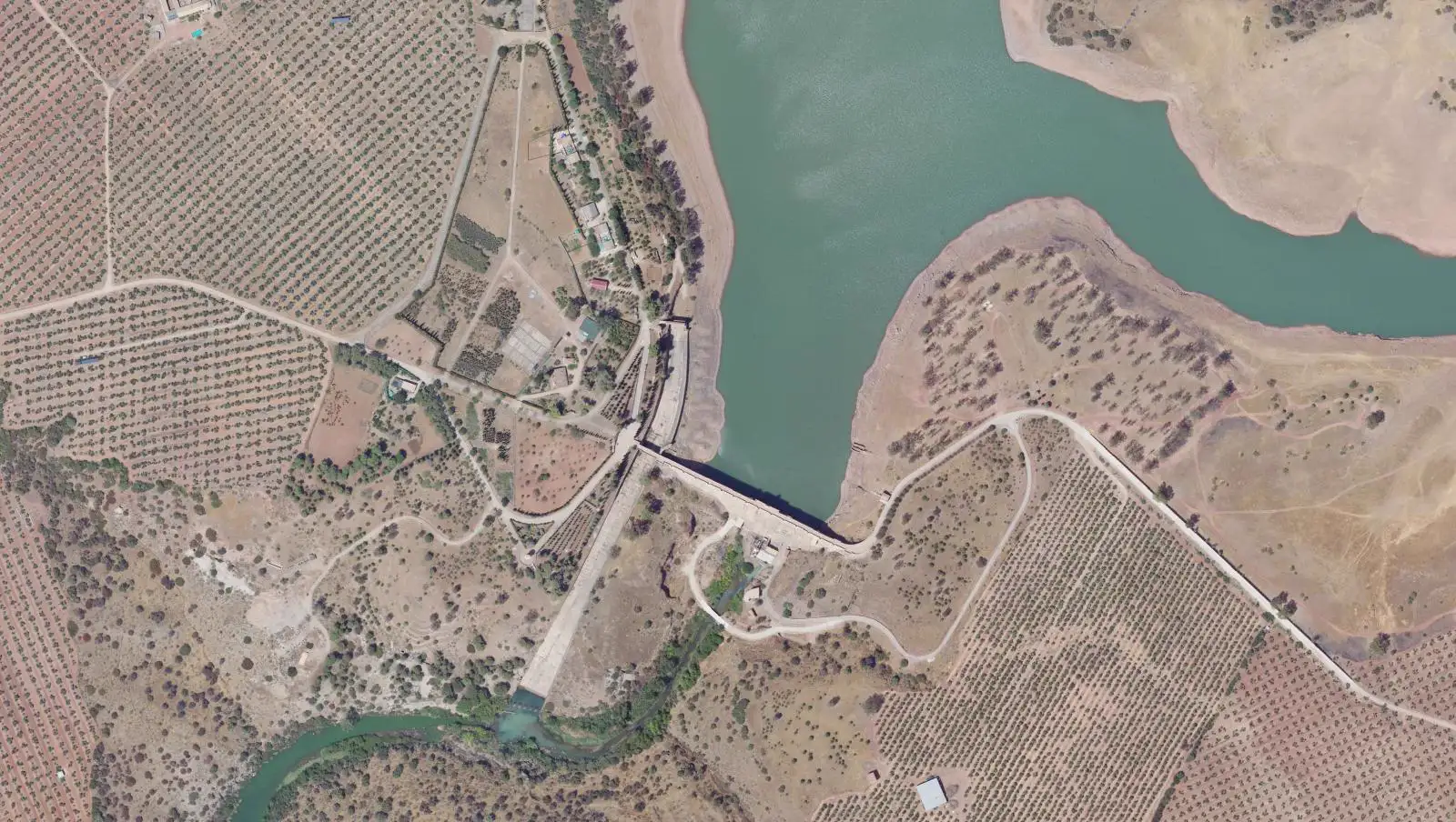 Vista aérea cenital de la presa del Guadalén: el muro cruza el valle, el embalse queda a la derecha y el canal del aliviadero baja en diagonal hasta el cauce del río, entre olivares
