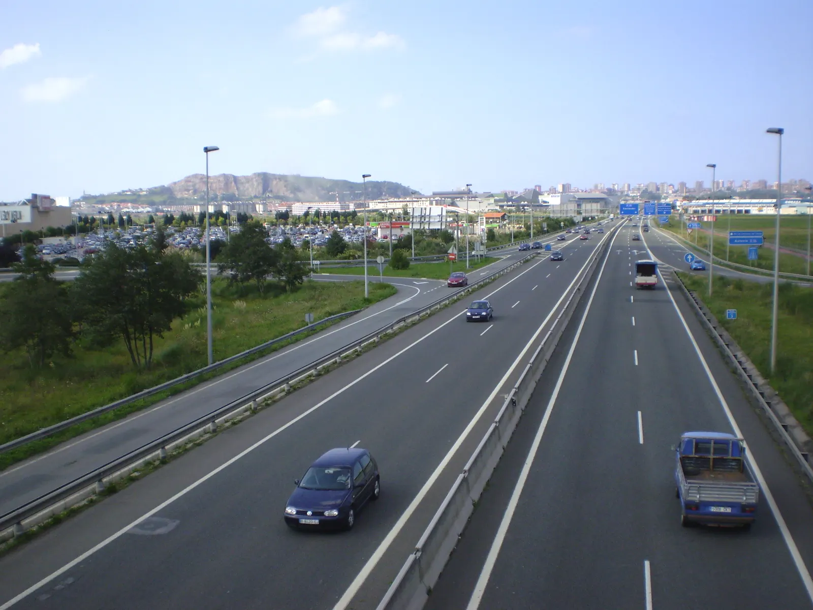 Vista elevada de la autovía S-10 con tráfico, aparcamientos y naves comerciales a la izquierda y el perfil urbano de Santander al fondo