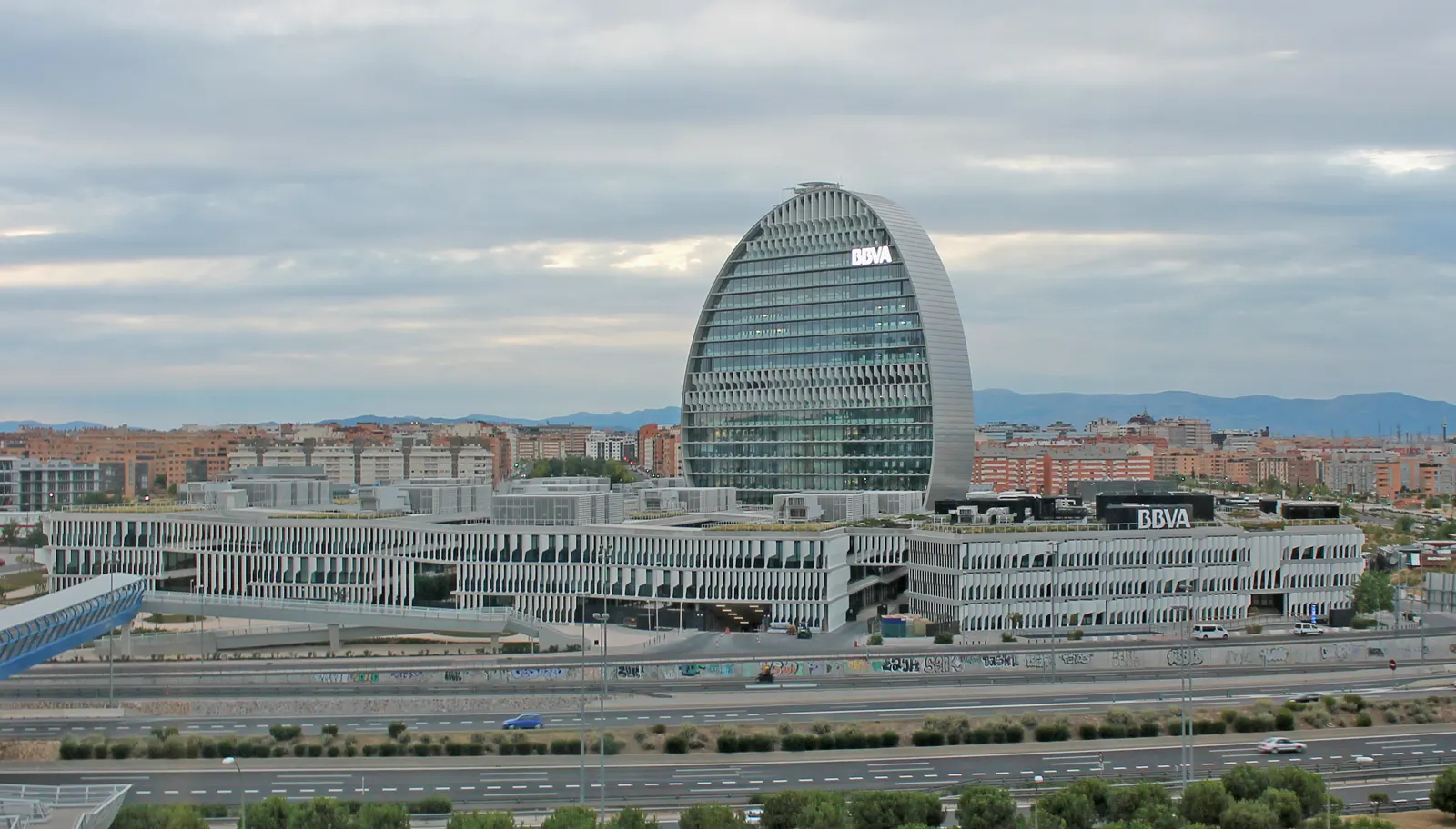 Vista general del complejo de oficinas Ciudad BBVA, en Madrid: edificios blancos de tres alturas con el rótulo BBVA y, detrás, una torre ovalada acristalada, con la ciudad y la autopista en primer plano.
