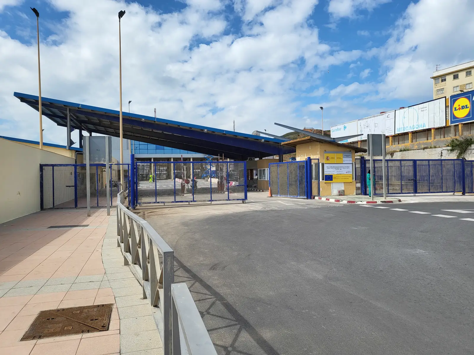 Puesto fronterizo de El Tarajal, en Ceuta, vacío: marquesina metálica azul sobre los carriles de paso, vallas metálicas cerradas, un cartel oficial con los escudos de España y de la Unión Europea y, al fondo, edificios y carteles de supermercados