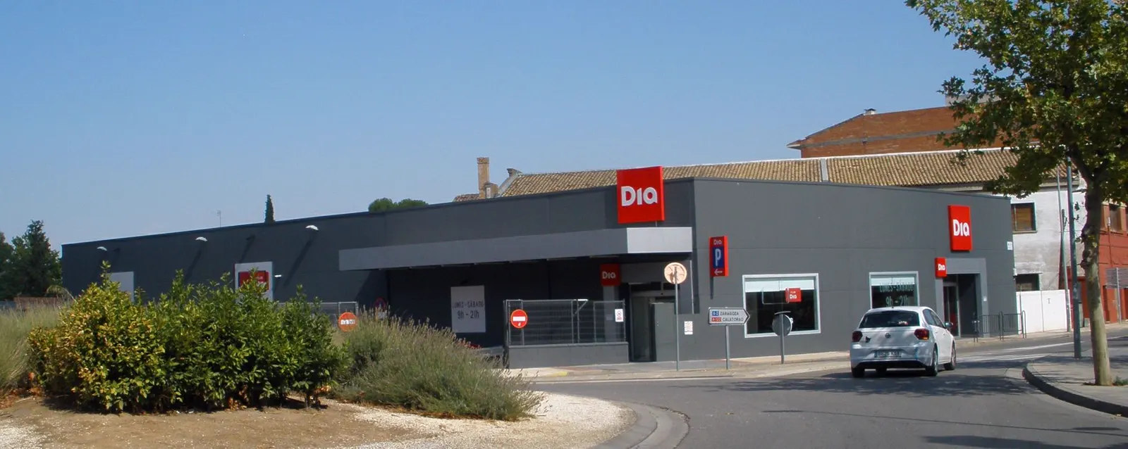 Supermercado Dia de una sola planta, con fachada gris y rótulos rojos con el logotipo de la cadena, junto a una rotonda con un coche blanco aparcado y arbustos en primer plano.