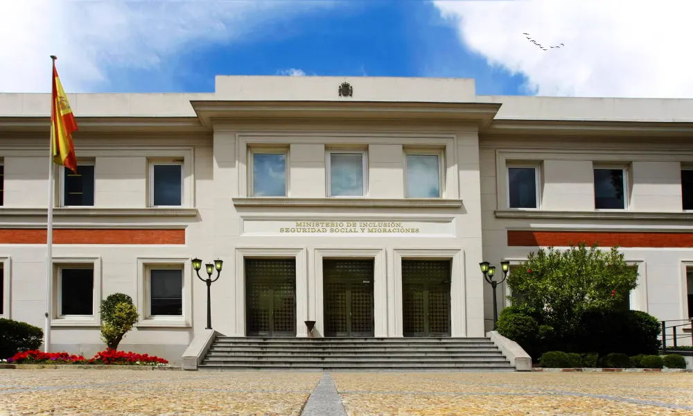 Fachada neoclásica de piedra clara de dos plantas con la inscripción «Ministerio de Inclusión, Seguridad Social y Migraciones» sobre la puerta principal, una bandera de España a la izquierda y una escalinata ante la entrada.