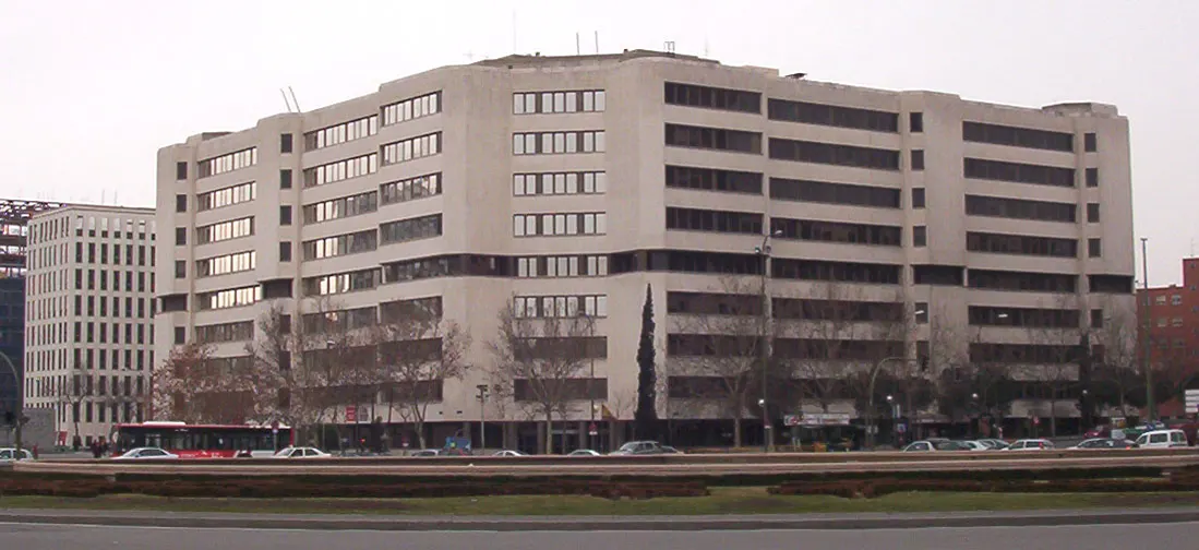 Fachada del edificio de los juzgados de la plaza de Castilla de Madrid, un bloque alargado de hormigón claro de nueve plantas con ventanas rectangulares en hilera; delante, una hilera de árboles sin hojas, coches aparcados y un autobús urbano, en un día nublado.