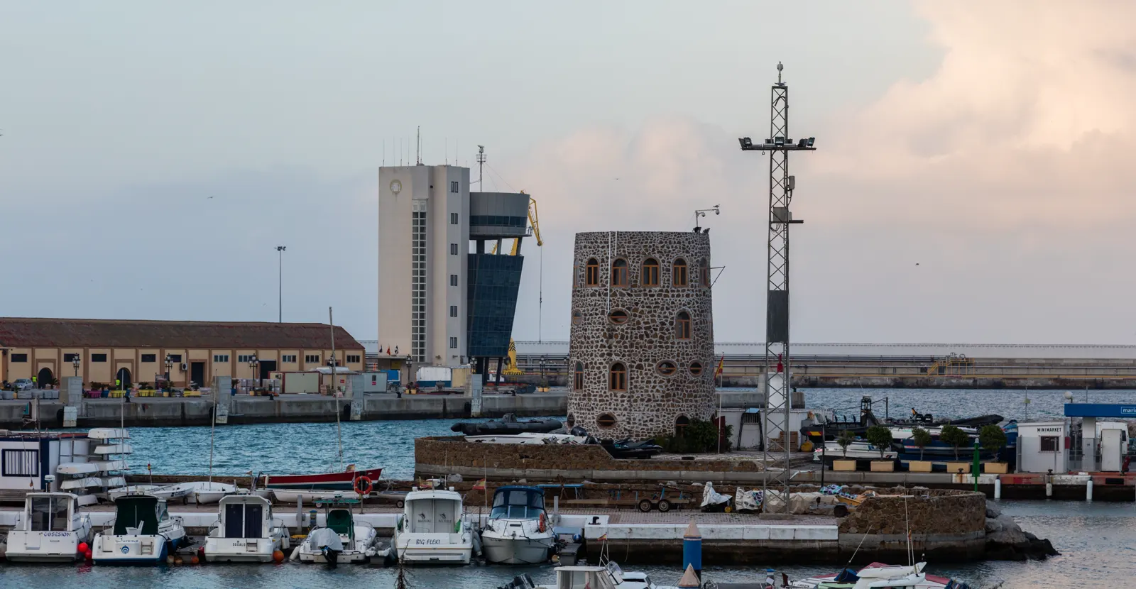 Vista del puerto de Ceuta al atardecer: pequeñas embarcaciones amarradas en primer plano, un torreón de piedra sobre el muelle, la torre blanca de control del puerto al fondo y el mar en calma bajo un cielo nublado.