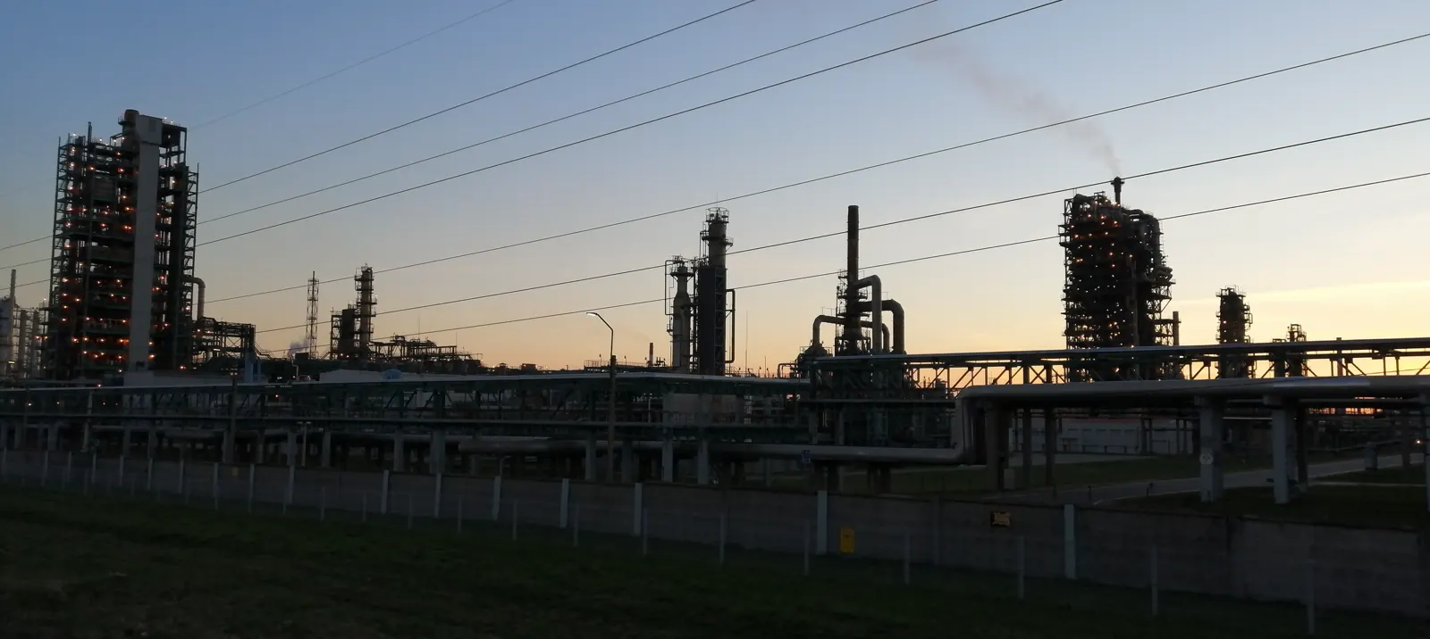 Perfil de una gran refinería de petróleo al atardecer, con torres de destilación iluminadas, chimeneas y tuberías elevadas tras una valla y un talud de hierba.