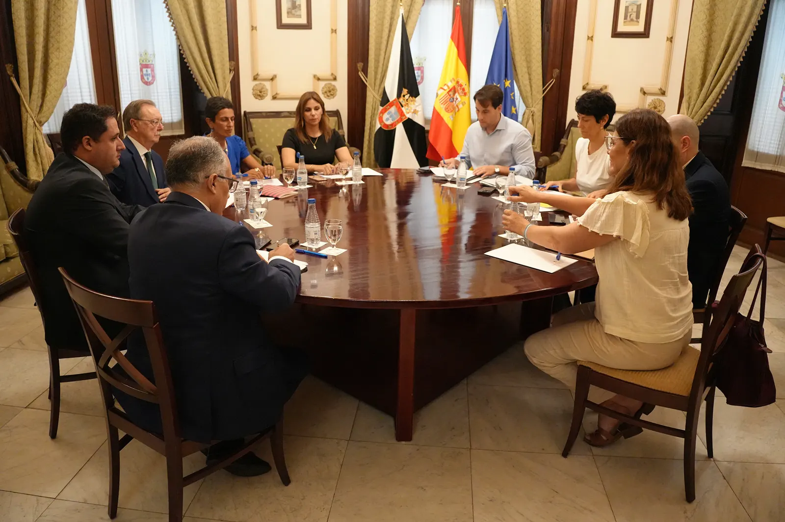 Diez personas sentadas alrededor de una gran mesa ovalada de madera en un salón institucional, con botellas de agua y carpetas ante ellas; al fondo, las banderas de Ceuta, España y la Unión Europea entre cortinas verdes.