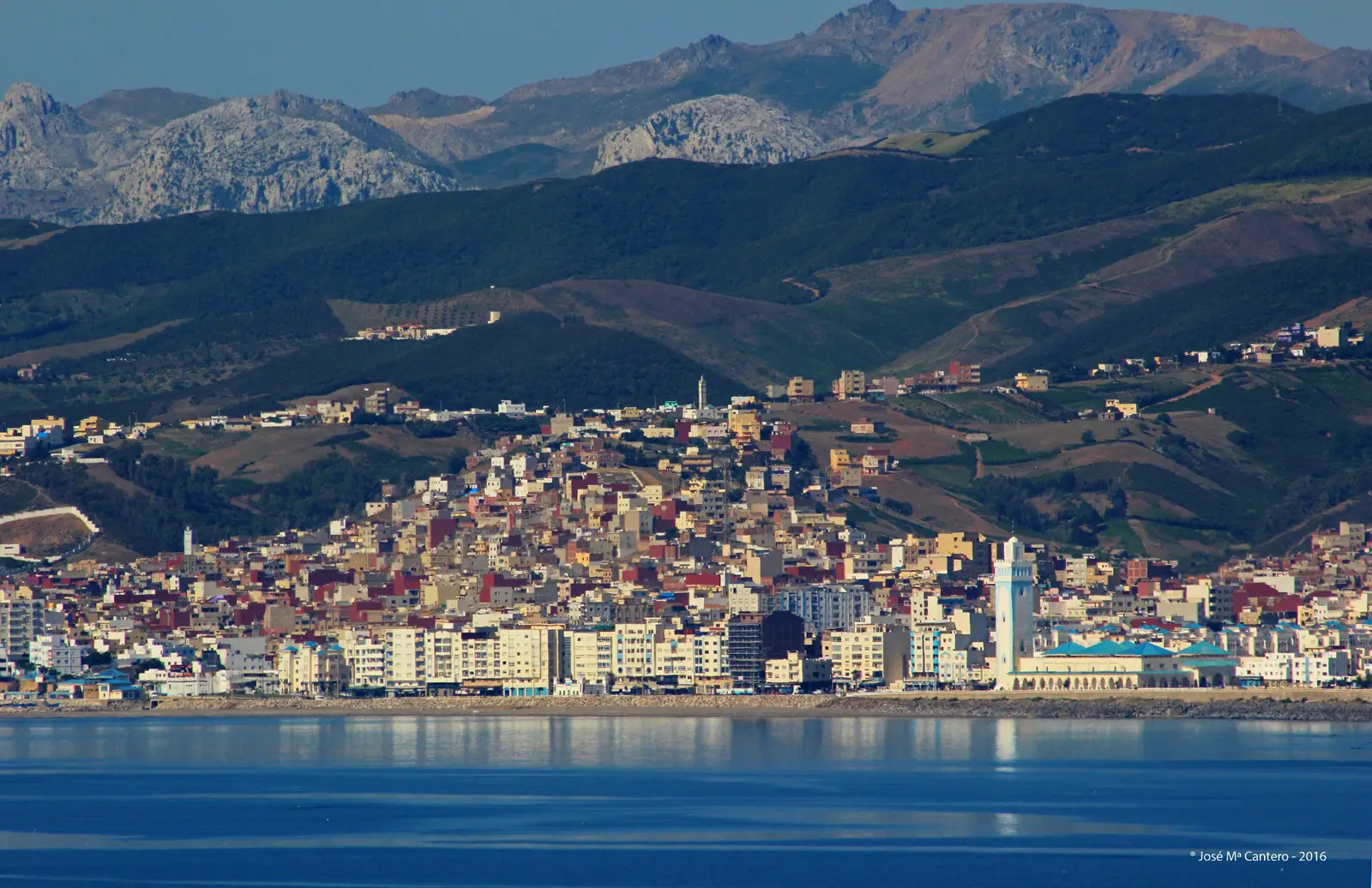 Vista de la ciudad marroquí de Fnideq (Castillejos) desde el mar: edificios blancos y ocres apiñados junto a una playa, una mezquita con minarete blanco y azul a la derecha y, al fondo, las montañas del Rif.