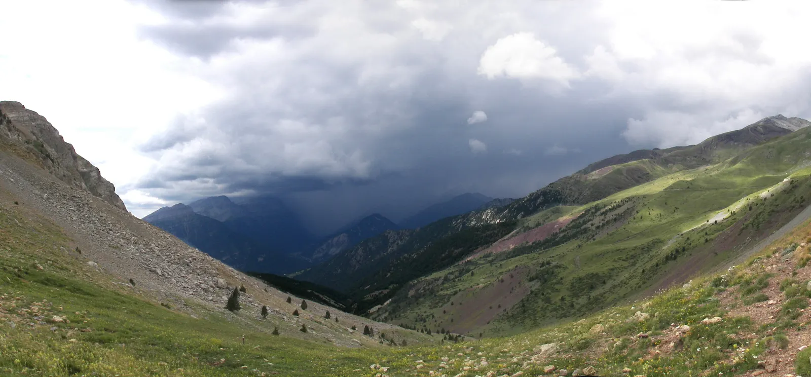 Vista desde un collado de montaña: laderas verdes y pedregales en primer plano y, al fondo del valle, una cortina oscura de lluvia que cae de un frente de nubes bajas sobre las cumbres del Pirineo oscense.