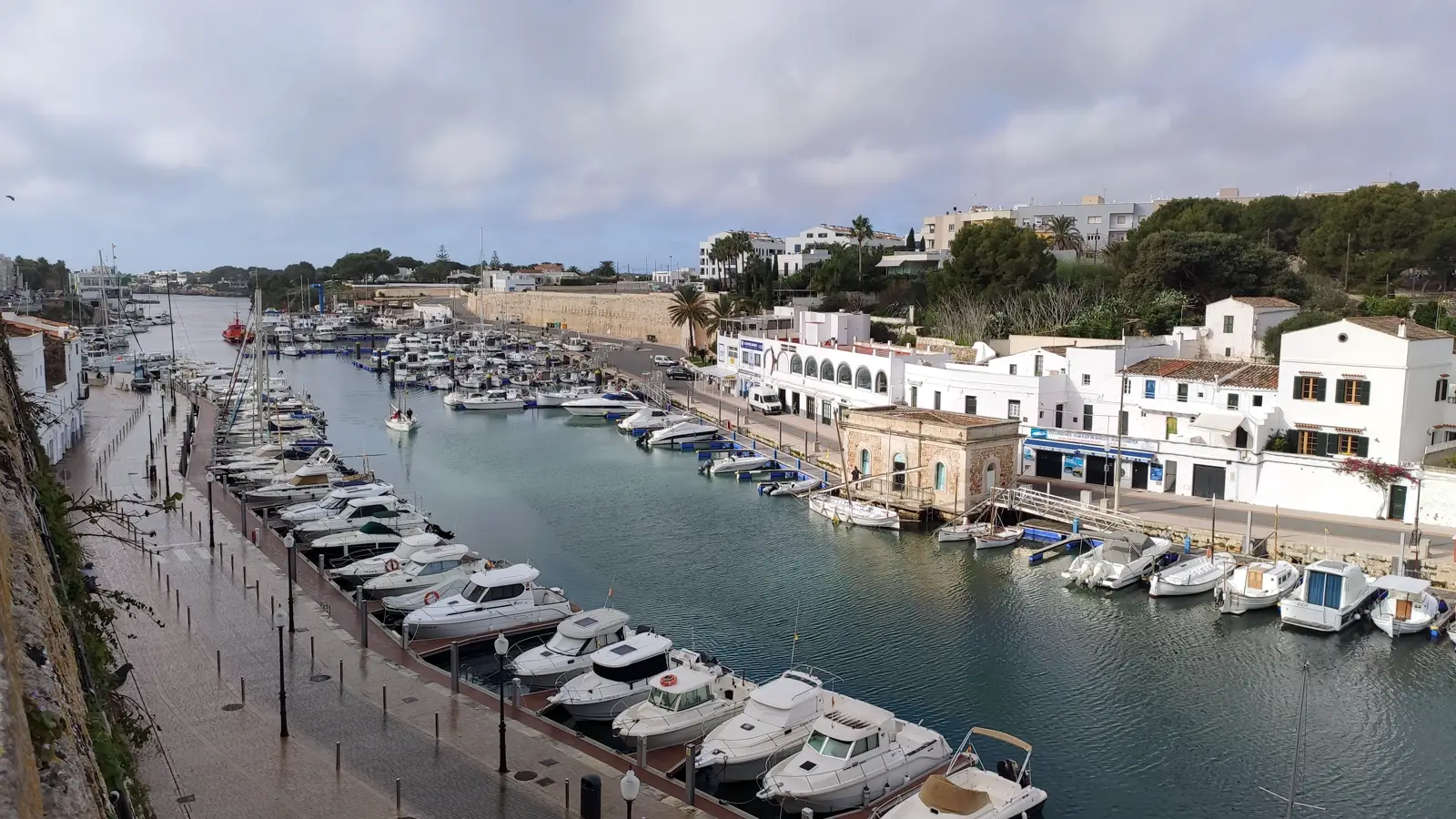Vista elevada del puerto de Ciutadella de Menorca en un día nublado: un canal de agua largo y estrecho encajado entre dos muelles, con decenas de embarcaciones de recreo amarradas en batería a ambos lados, un paseo peatonal mojado a la izquierda y casas blancas de dos plantas a la derecha.