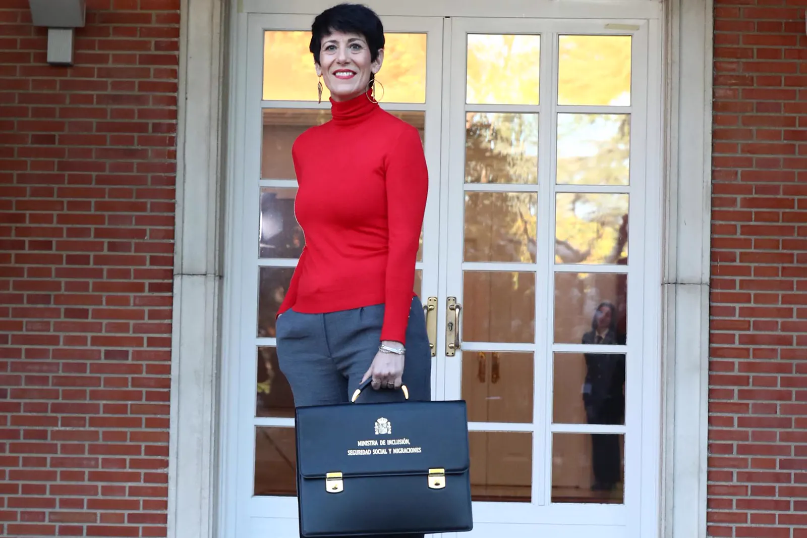 Elma Saiz, de jersey rojo de cuello alto y pantalón gris, sostiene con la mano derecha la cartera ministerial con el escudo de España y la leyenda «Ministra de Inclusión, Seguridad Social y Migraciones», ante una puerta acristalada blanca de un edificio de ladrillo.