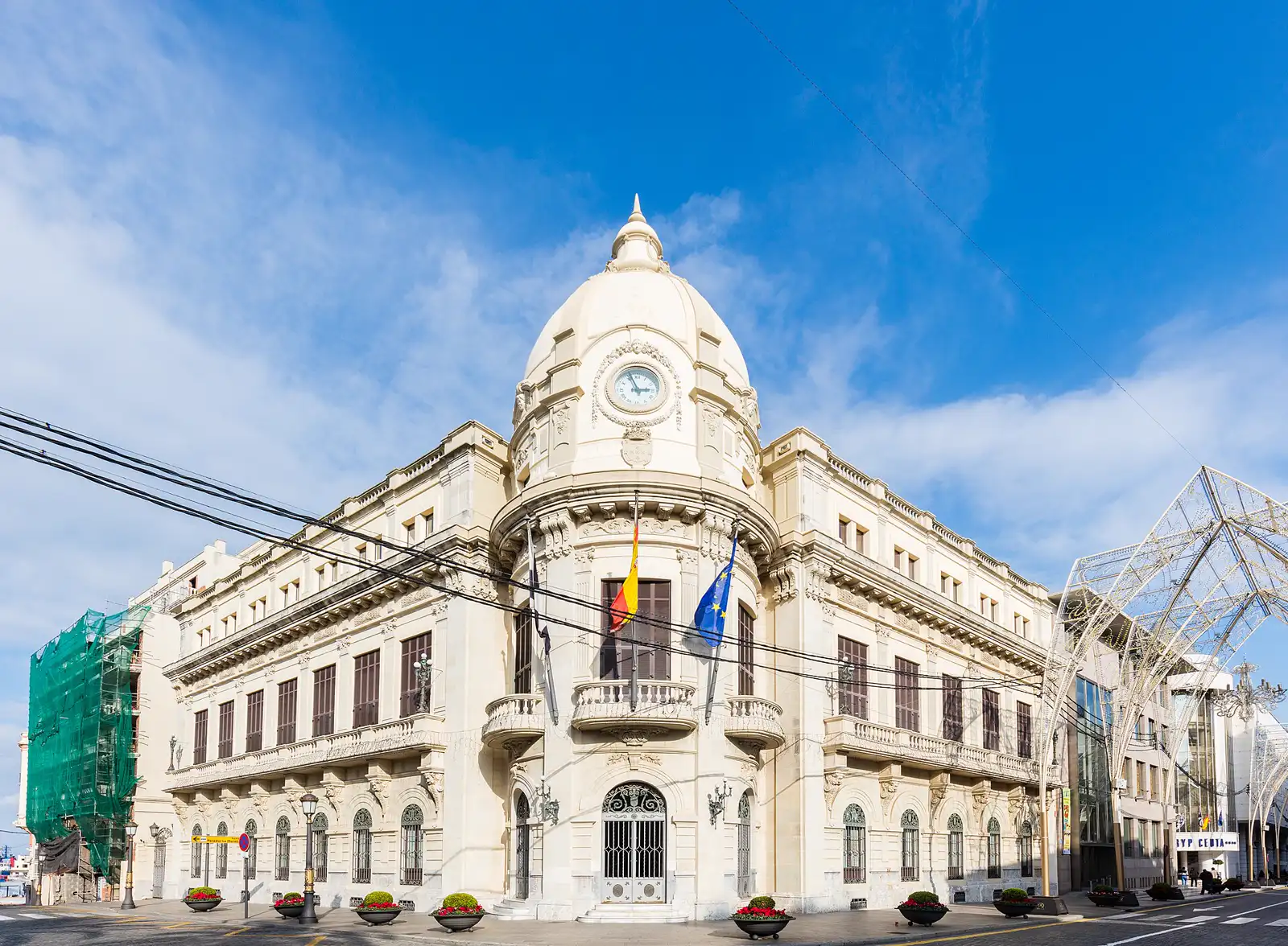 Edificio institucional de piedra clara con cúpula y reloj en la esquina, visto desde la calle bajo un cielo azul. En el balcón principal ondean las banderas de Ceuta, España y la Unión Europea; a pie de calle hay maceteros con flores rojas y setos recortados.