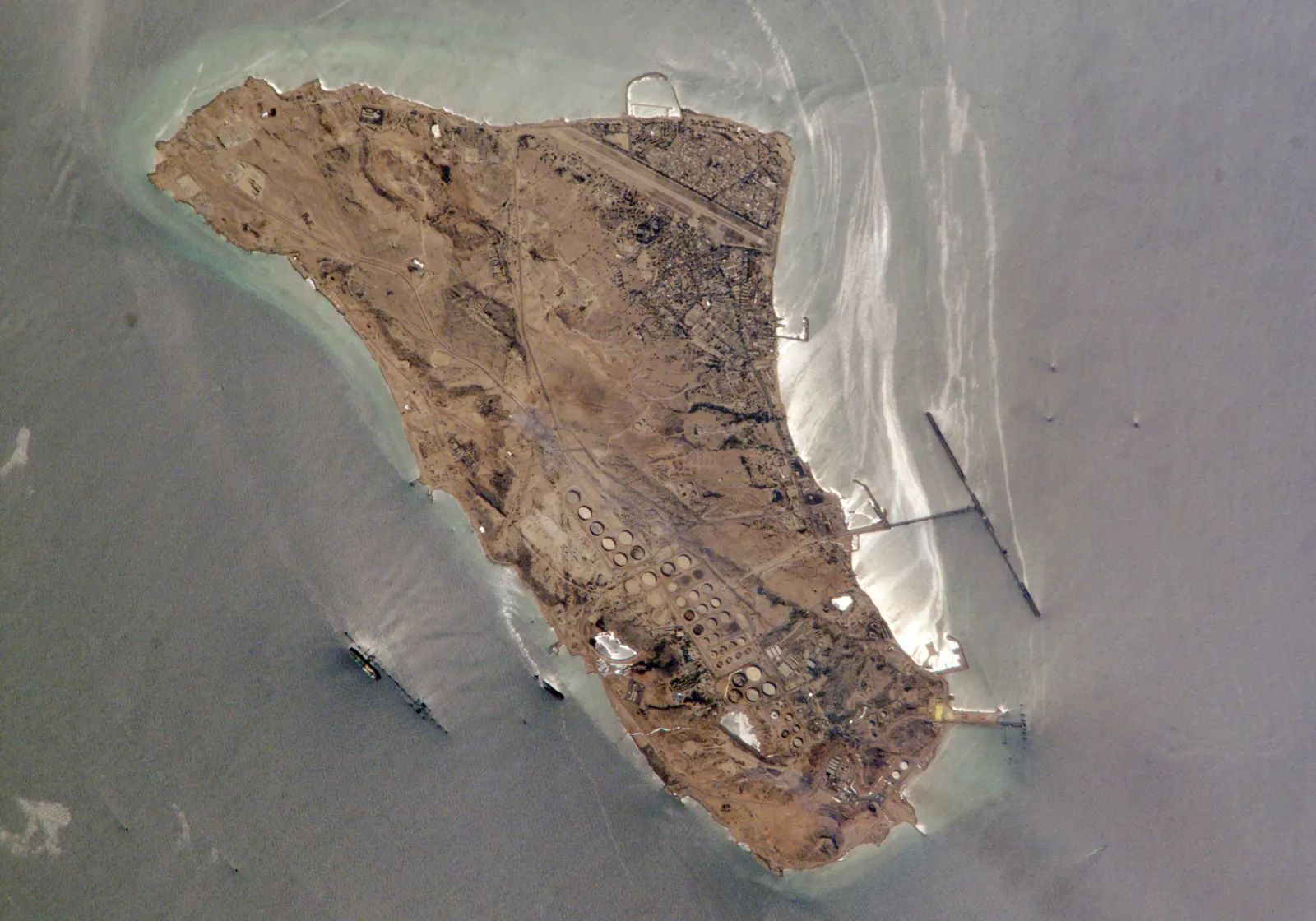 Vista cenital desde el espacio de la isla de Jarg, en el golfo Pérsico: una isla alargada de terreno árido con una pista de aterrizaje, un núcleo urbano al norte, decenas de depósitos circulares de crudo en el sur y un largo muelle en forma de T que se adentra en el mar, con petroleros fondeados alrededor.