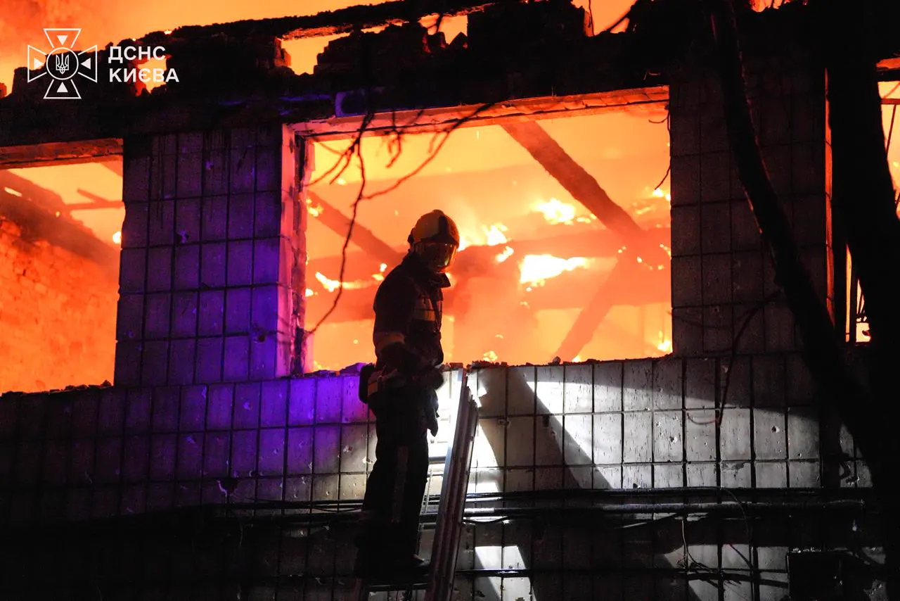 Un bombero con casco y equipo de protección, de pie sobre una escalera apoyada en un muro acribillado, recortado contra el interior en llamas de un edificio sin techo, de noche.
