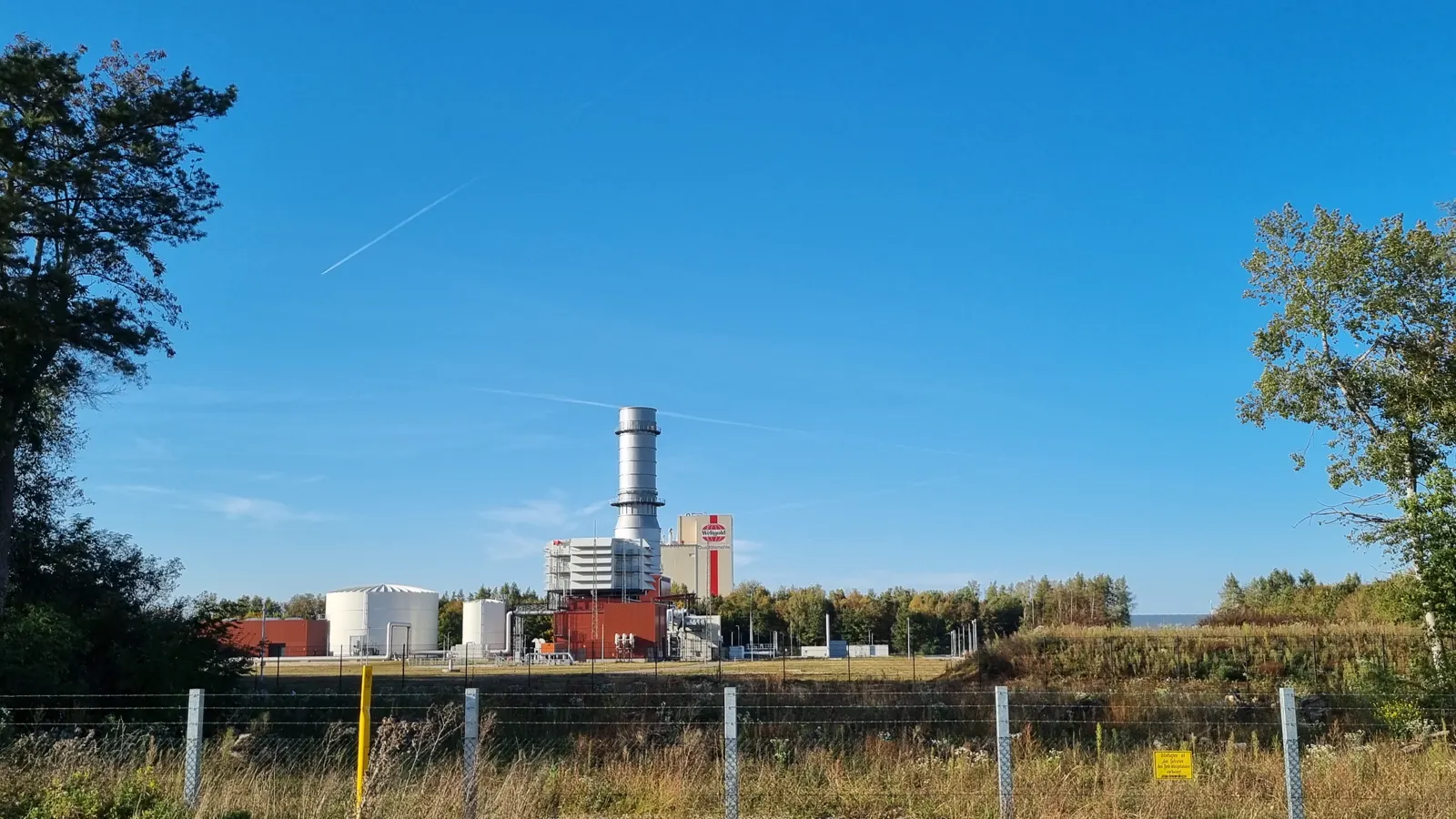 Central de gas con una chimenea metálica alta y naves rojas y blancas al fondo de un prado, tras una valla metálica, bajo un cielo azul despejado.