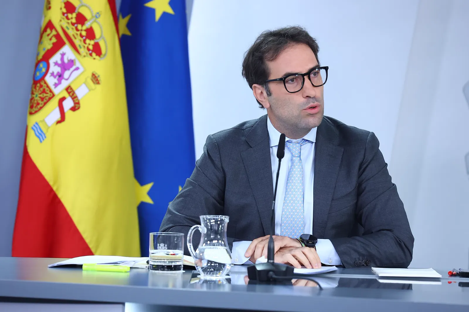 Un hombre de mediana edad con gafas de pasta negra, traje gris y corbata azul claro habla ante un micrófono sentado a una mesa con una jarra y un vaso de agua; detrás, las banderas de España y de la Unión Europea sobre un panel blanco.
