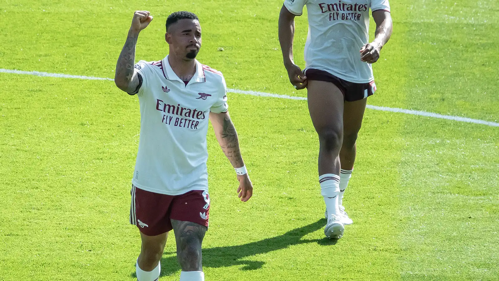 Gabriel Jesus celebra un gol con el puño en alto, vestido con la equipación blanca y granate del Arsenal, mientras un compañero se acerca corriendo por el césped.