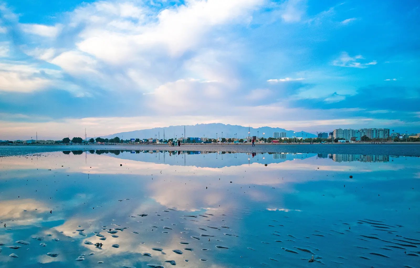 Vista de la ciudad iraní de Bandar Abbas desde la orilla al atardecer: una lámina de agua en la marea baja refleja el cielo nublado, y al fondo se alinean edificios de viviendas y palmeras con una cordillera árida recortada en el horizonte.