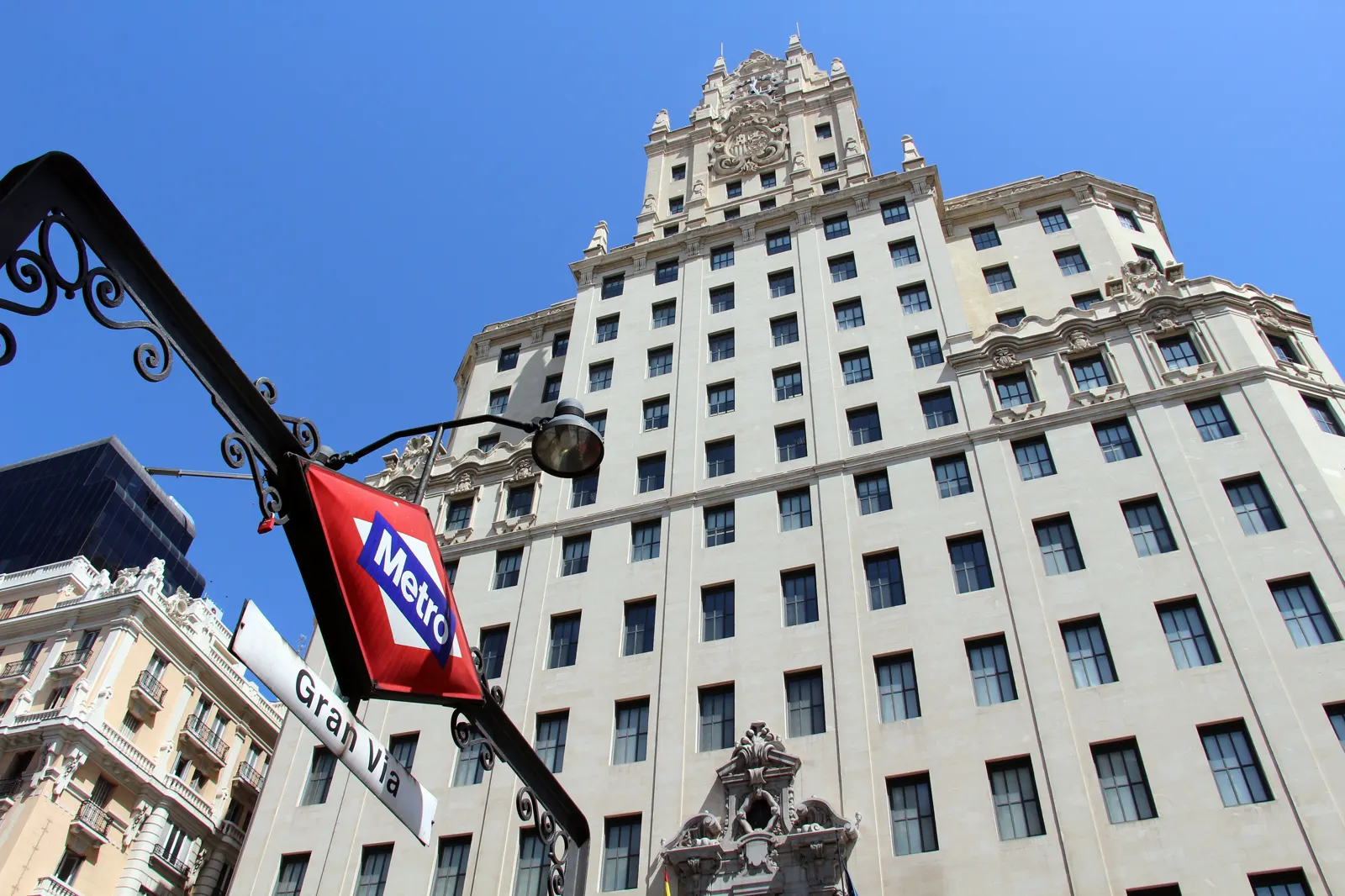 Fachada del edificio de Telefónica en la Gran Vía de Madrid vista en contrapicado, con su remate neobarroco recortado sobre un cielo azul y, en primer plano, un farol con el rótulo del metro de Gran Vía.