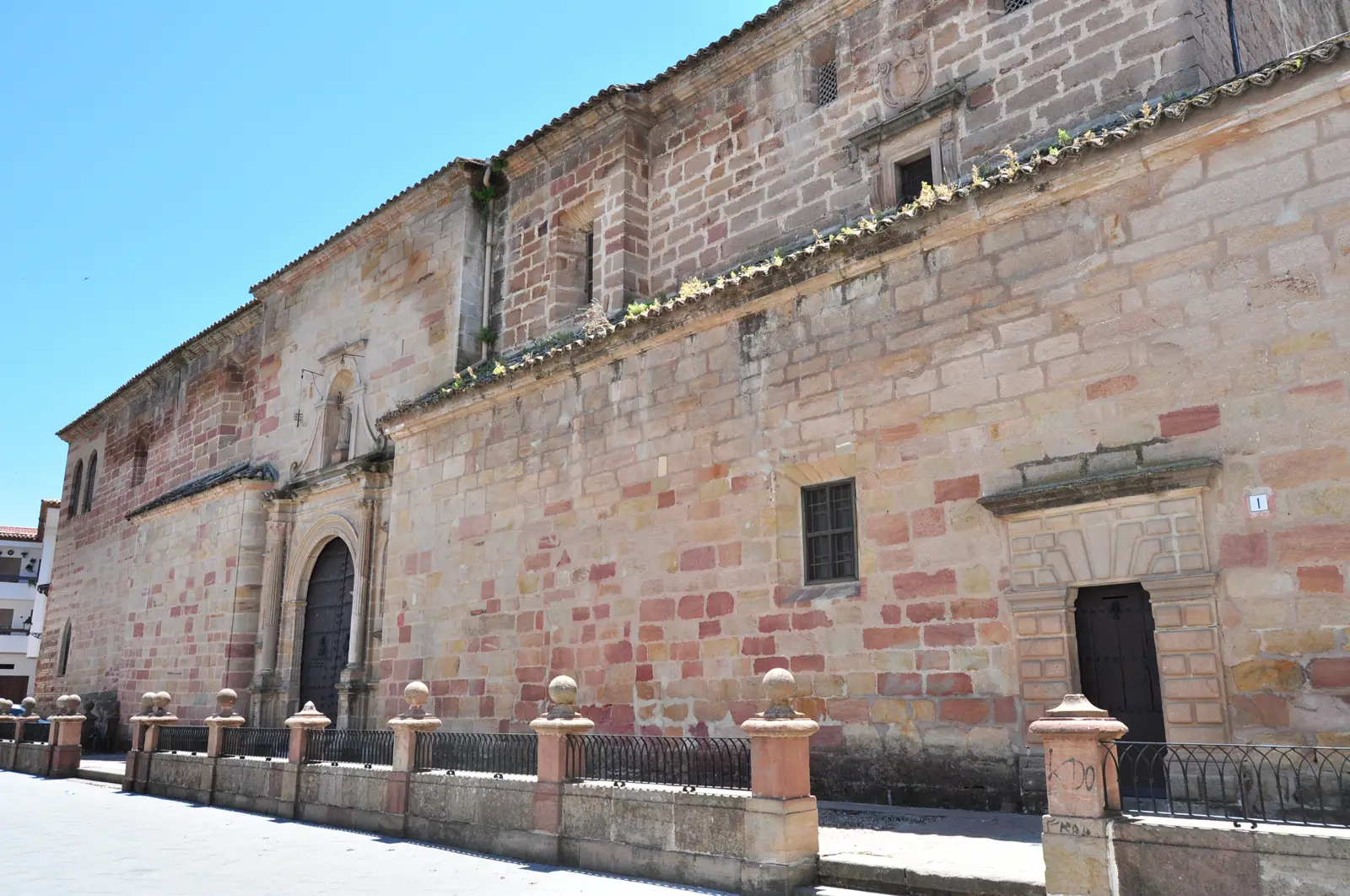 Muro lateral de sillería rosada de la iglesia de Santa María la Mayor de Andújar bajo un sol vertical: la piedra recibe la luz de plano, las sombras de las bolas del pretil son cortas y redondas, la acera aparece vacía y el cielo, de un azul intenso, no tiene una sola nube.