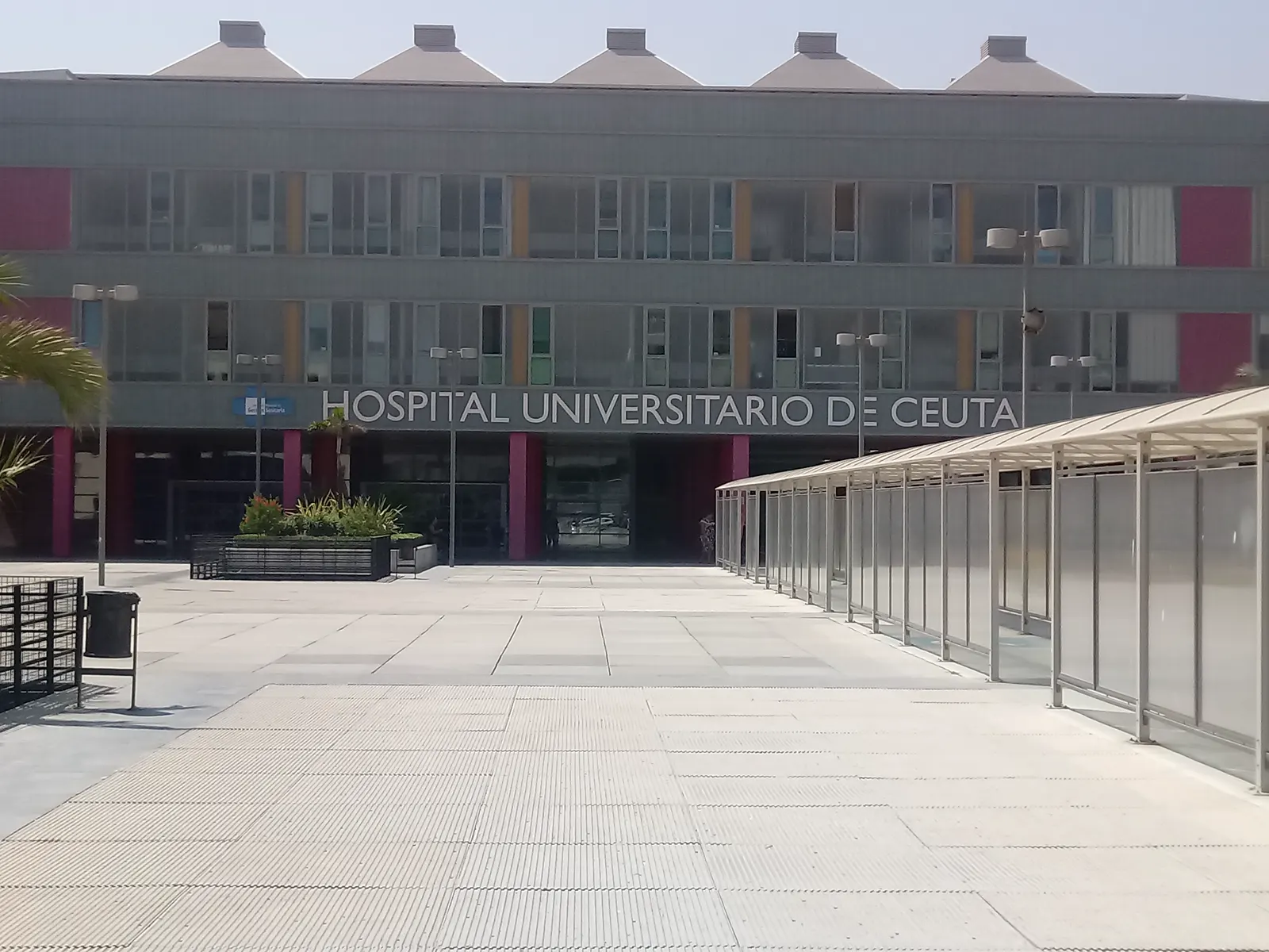 Fachada acristalada de tres plantas con rótulo «Hospital Universitario de Ceuta» sobre la entrada principal, con pilares rojos, una explanada peatonal vacía en primer plano y una marquesina metálica a la derecha.
