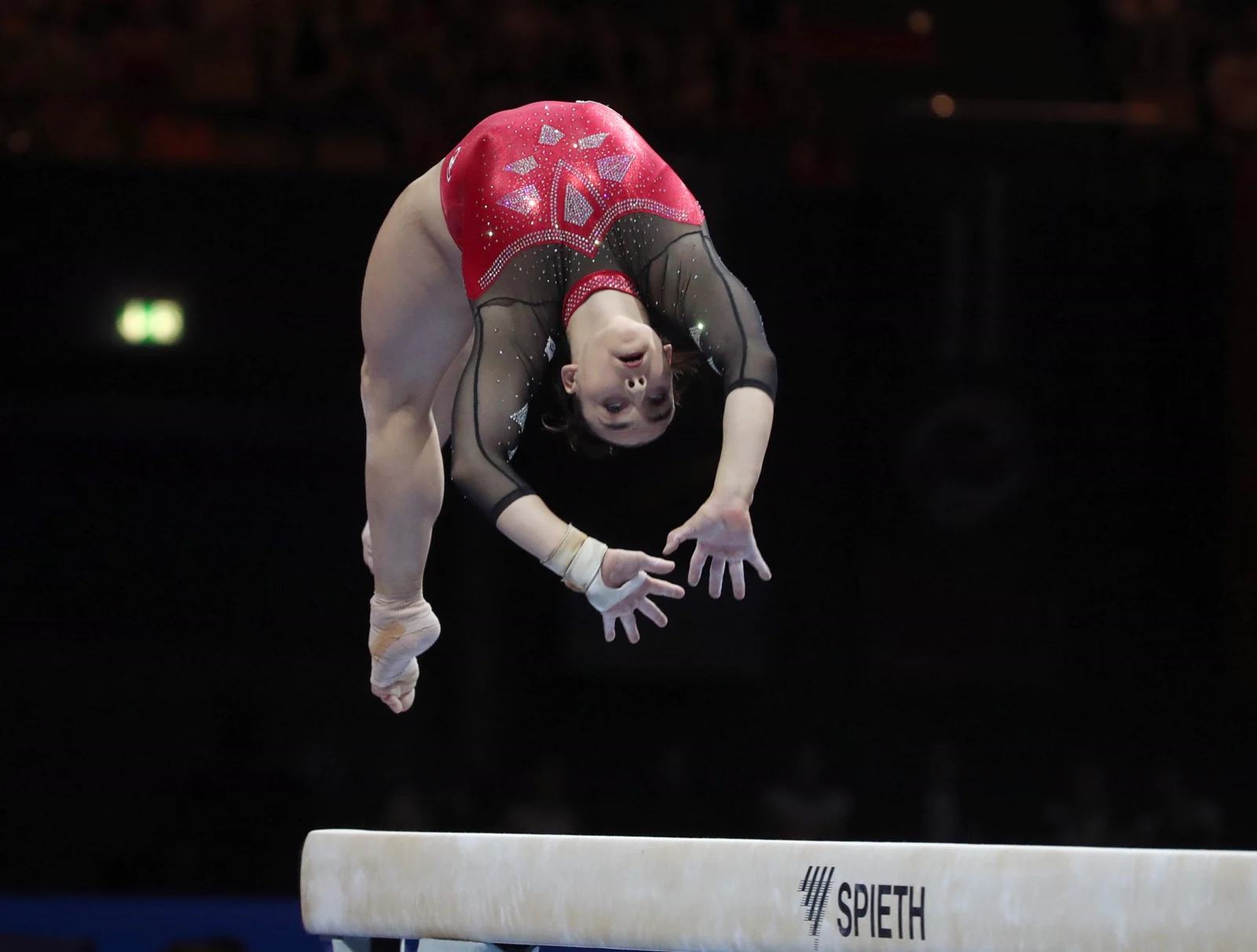 Una gimnasta con maillot rojo y negro, en pleno salto acrobático con el cuerpo arqueado y las manos buscando el apoyo, sobre una barra de equilibrio.
