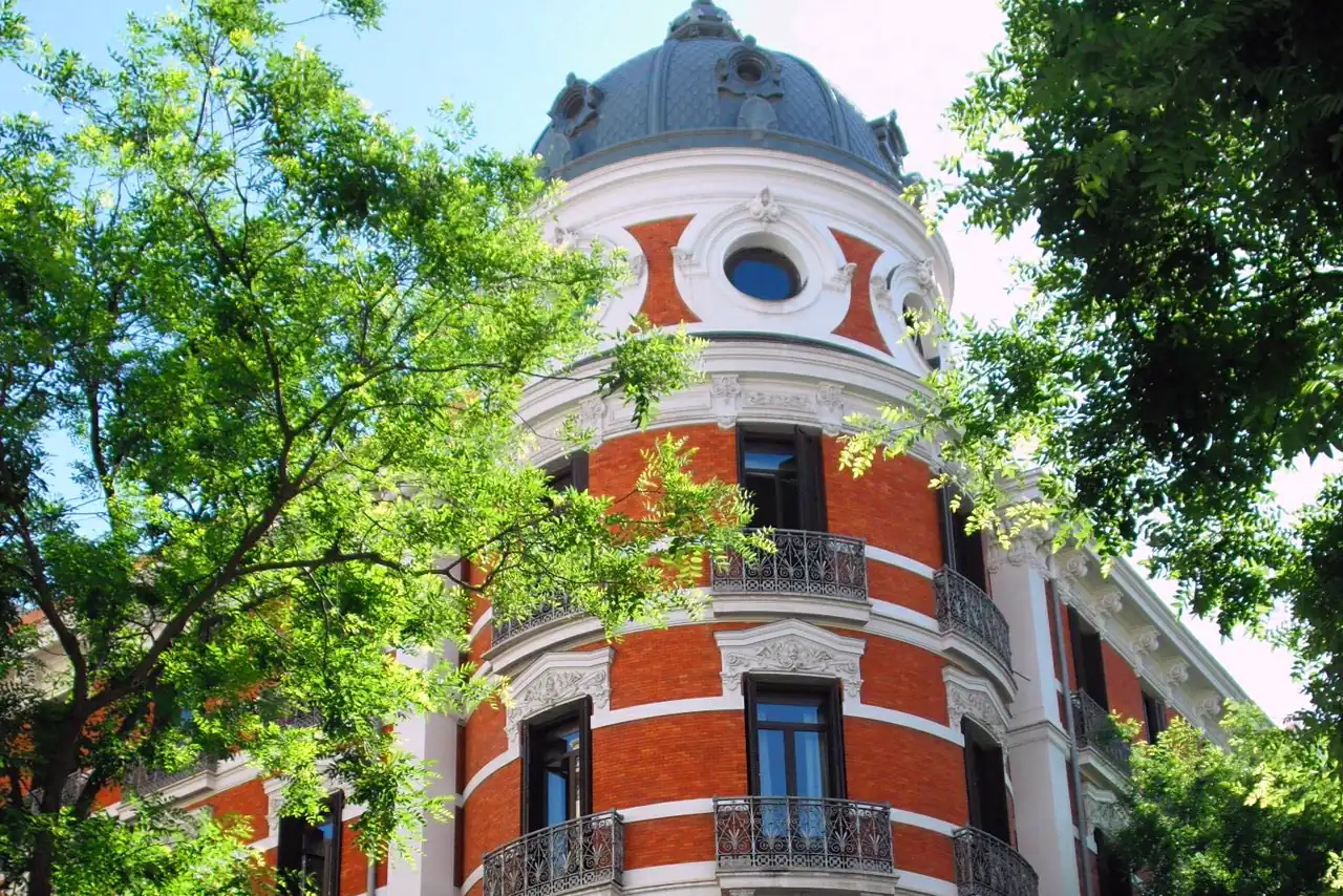Esquina de un edificio de ladrillo rojo y molduras blancas rematada por una cúpula de pizarra con óculos y balcones de hierro forjado, vista desde la acera entre las ramas de dos árboles.