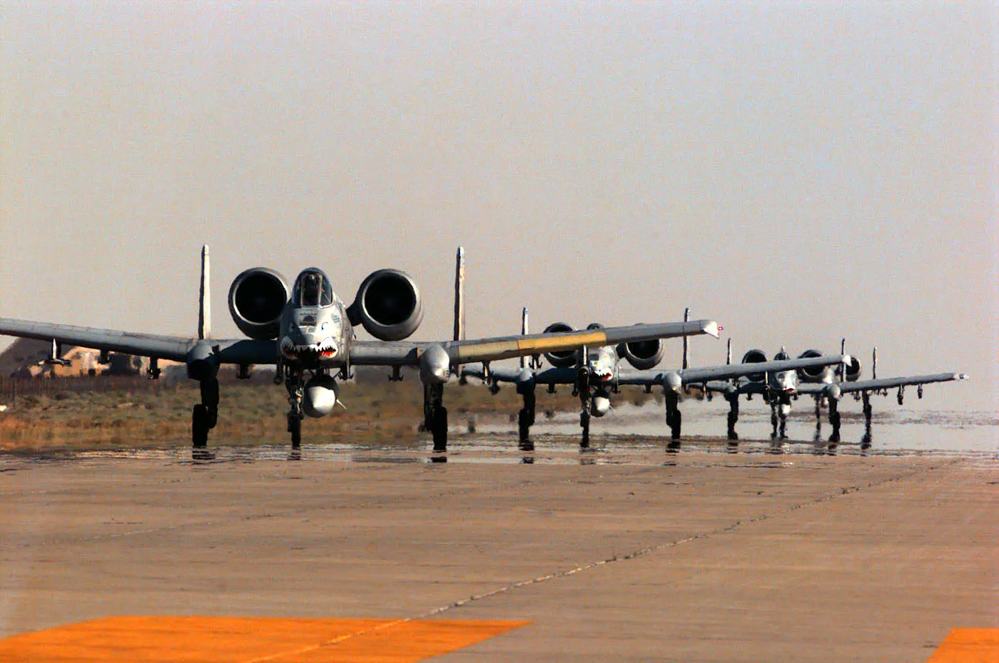 Cuatro aviones de ataque A-10 Thunderbolt II en fila sobre la pista de una base aérea en el desierto, vistos de frente y con fauces de tiburón pintadas en el morro; el asfalto reverbera por el calor y el horizonte se difumina en una bruma gris.