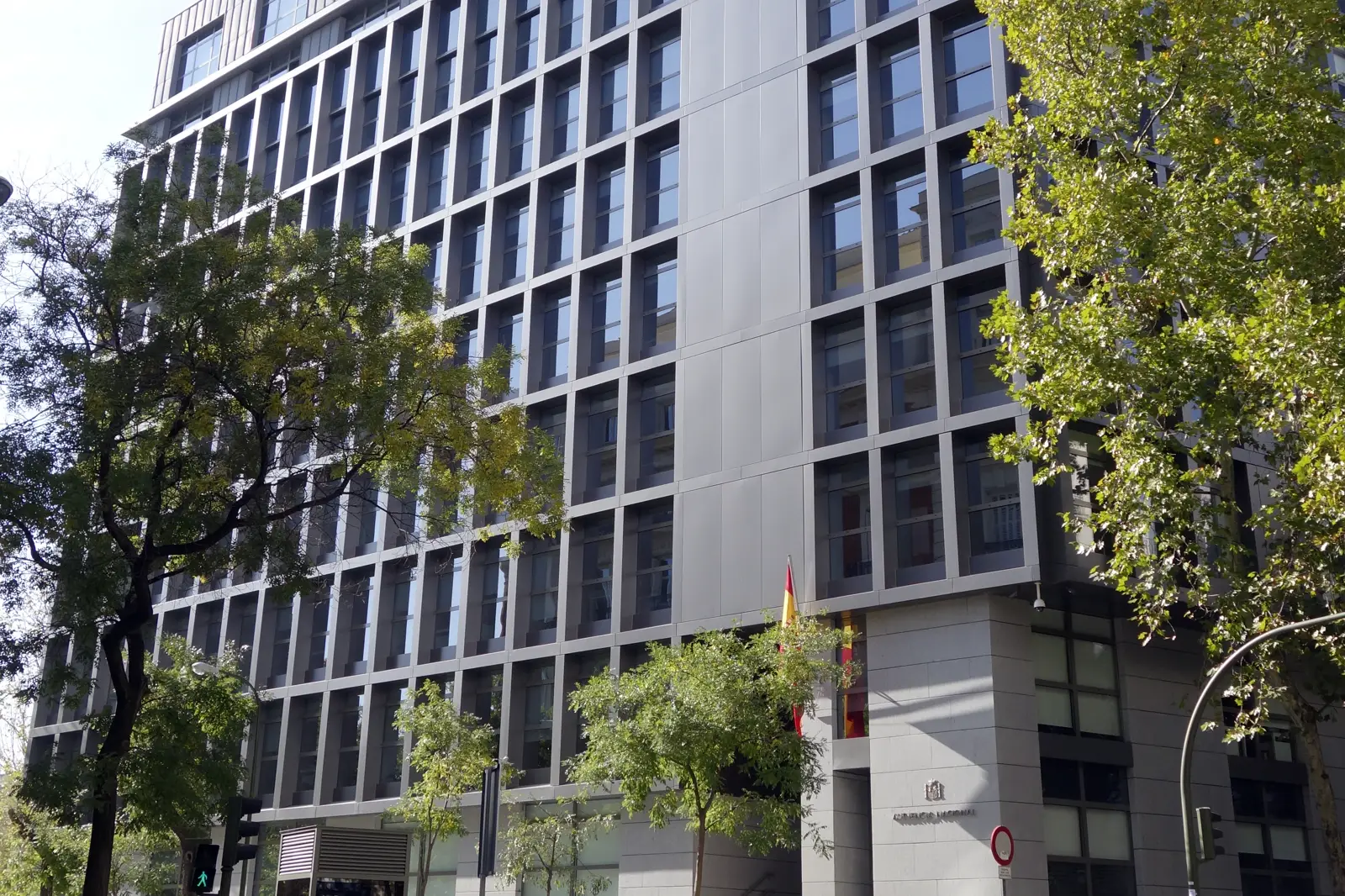 Fachada de la sede de la Audiencia Nacional en Madrid, un edificio moderno de cristal y hormigón con la bandera de España sobre la entrada, entre árboles