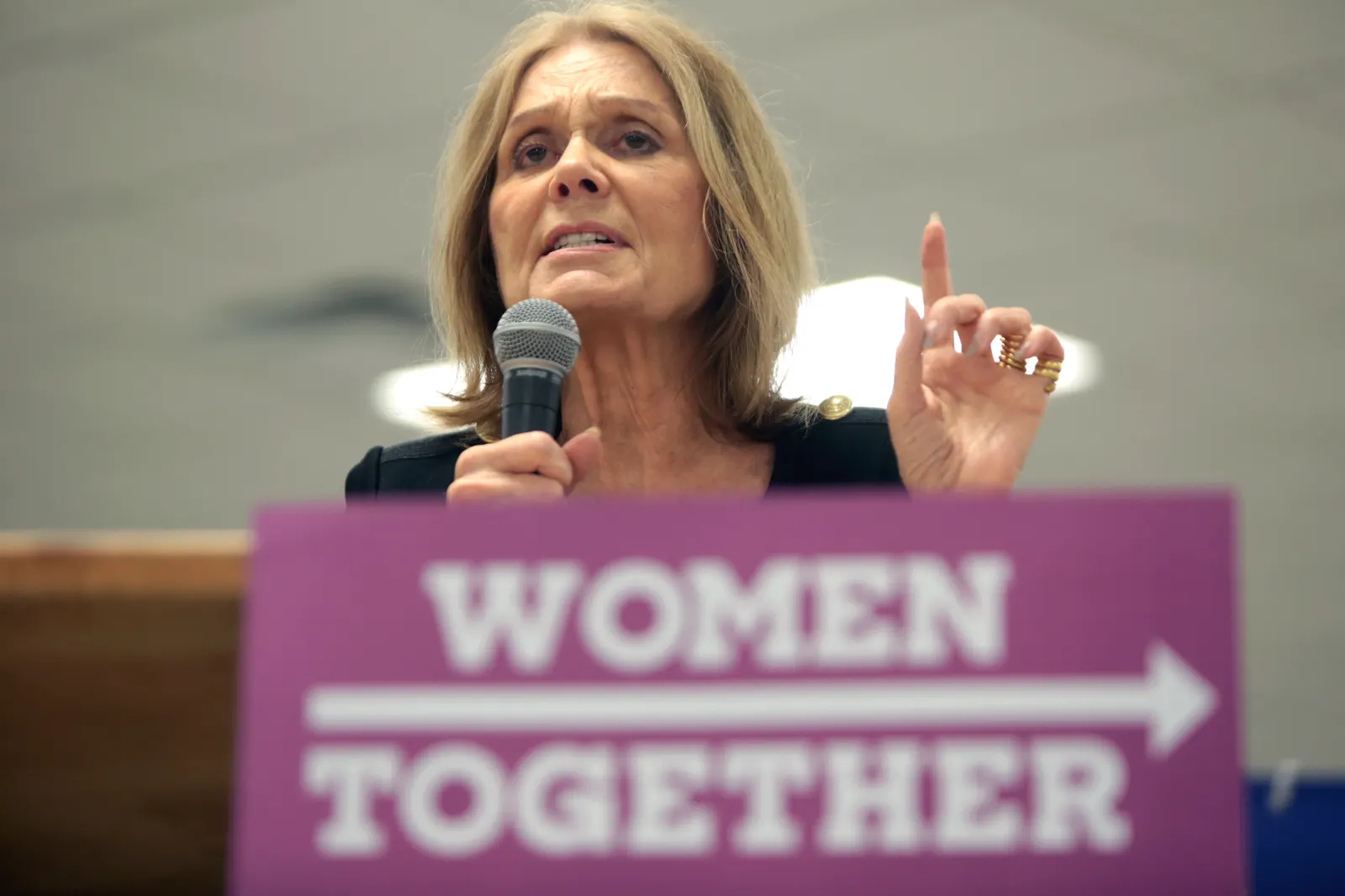 Gloria Steinem habla con un micrófono en la mano y el índice levantado tras un atril morado con el lema «Women Together»