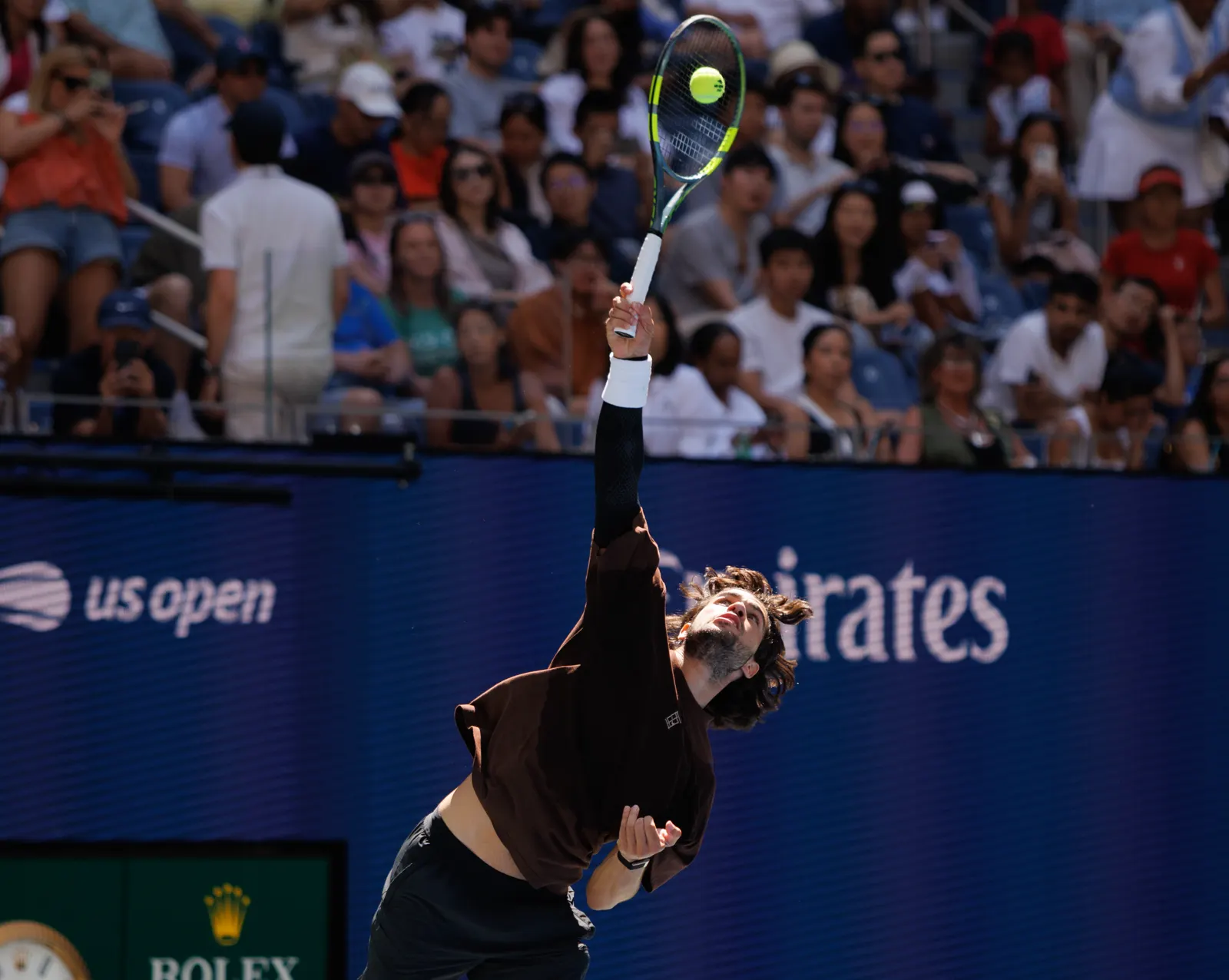 Un tenista con camiseta oscura salta para golpear un saque; la pelota está aún sobre el cordaje de la raqueta, en lo alto. Detrás, el panel azul de la pista con el rótulo «us open» y la grada llena de público.