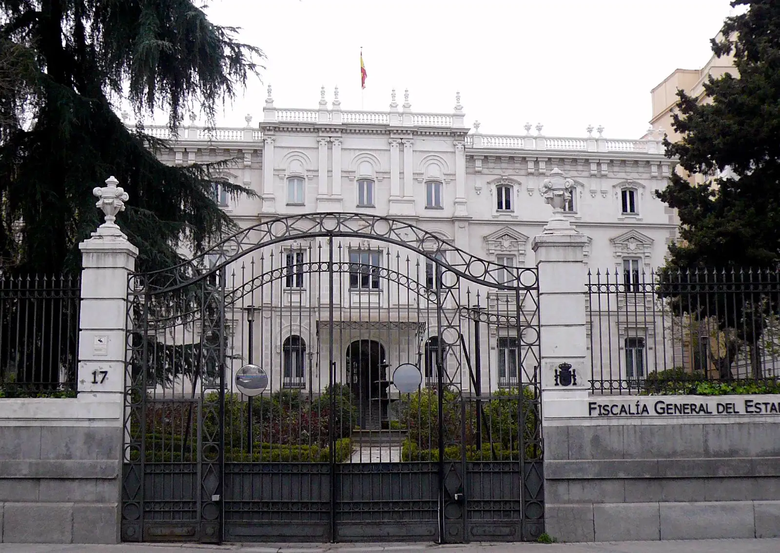 Fachada blanca de un palacio de tres plantas con balaustrada y bandera de España en la cubierta, vista desde la calle tras una verja de hierro negro; en el muro de la derecha, el escudo de España y el rótulo «Fiscalía General del Estado»; en el pilar de la izquierda, el número 17.