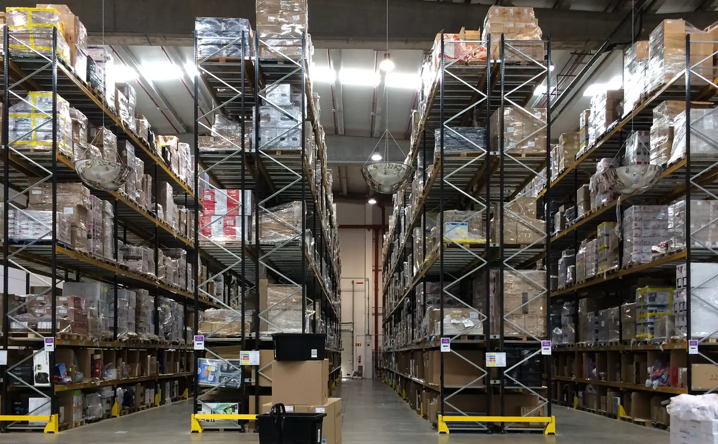 Pasillo de un centro logístico entre dos estanterías metálicas de gran altura repletas de palés con cajas envueltas en plástico
