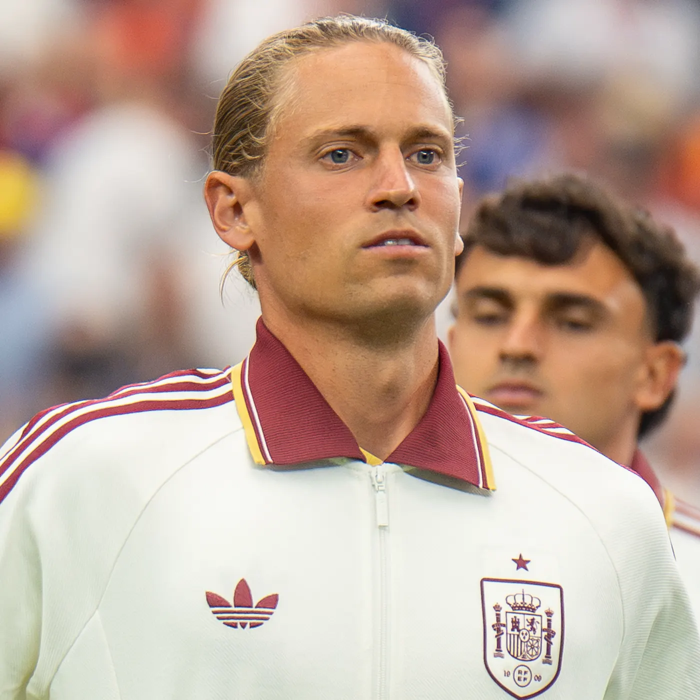 Marcos Llorente, rubio y con el pelo recogido, vestido con la chaqueta blanca de la selección española —cuello granate, escudo de la Real Federación Española de Fútbol y una estrella sobre él en el pecho—, mira serio hacia un lado; detrás, desenfocado, otro jugador