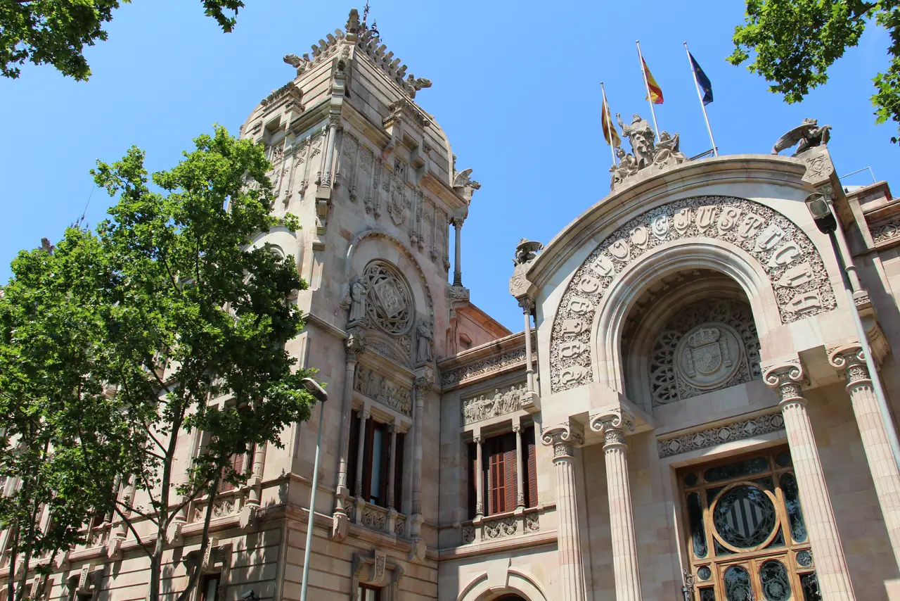 Fachada modernista del Palau de Justícia de Barcelona vista en contrapicado: gran arco de piedra con la inscripción «Palau de Justícia», torre lateral y tres banderas ondeando sobre la cornisa bajo un cielo azul.