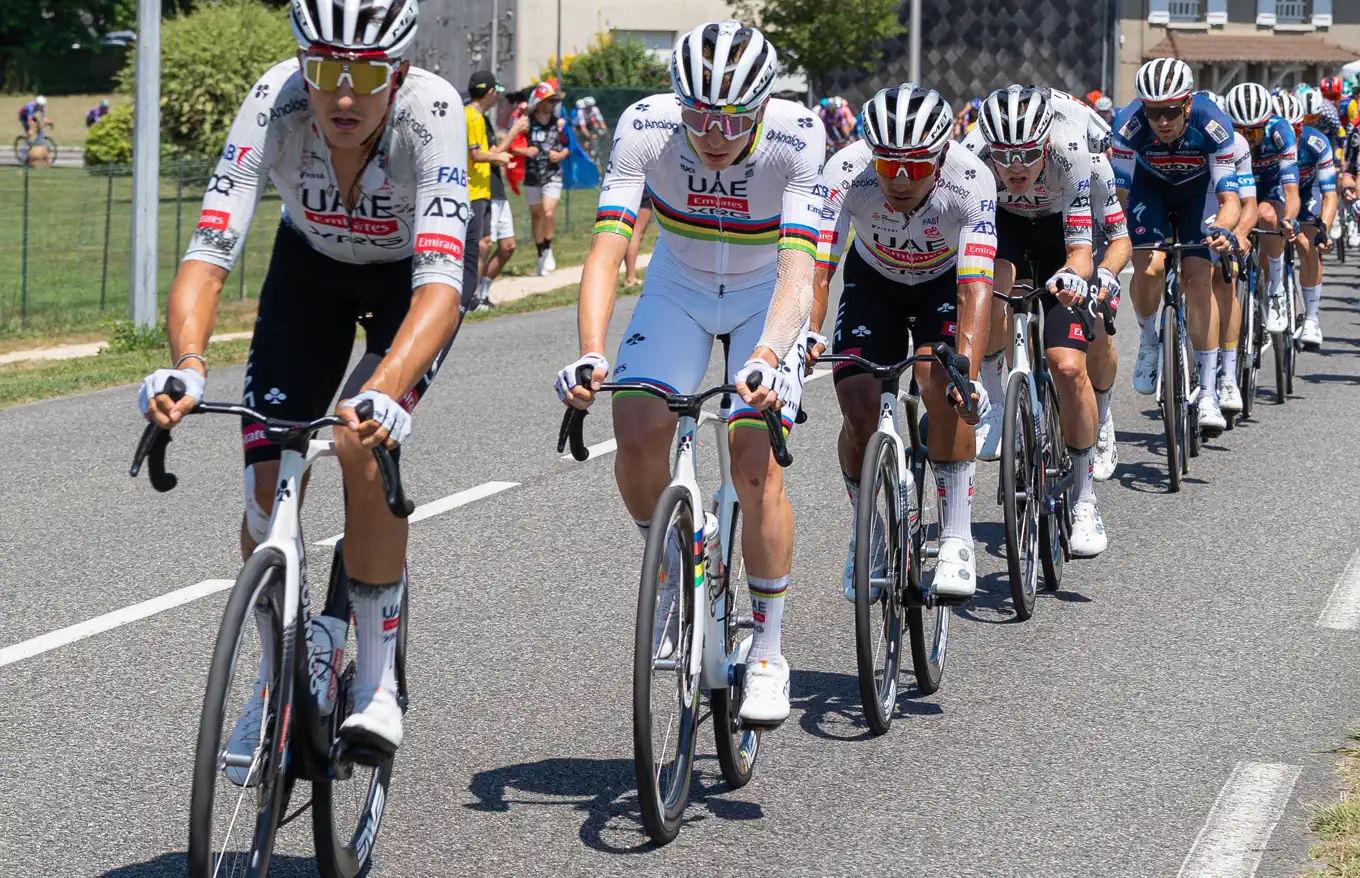 Un grupo de ciclistas rueda en fila por una carretera; el segundo por la izquierda viste el maillot blanco con franjas de arcoíris de campeón del mundo y lleva el logotipo de UAE Emirates en el pecho, flanqueado por compañeros con la equipación blanca y negra del mismo equipo.