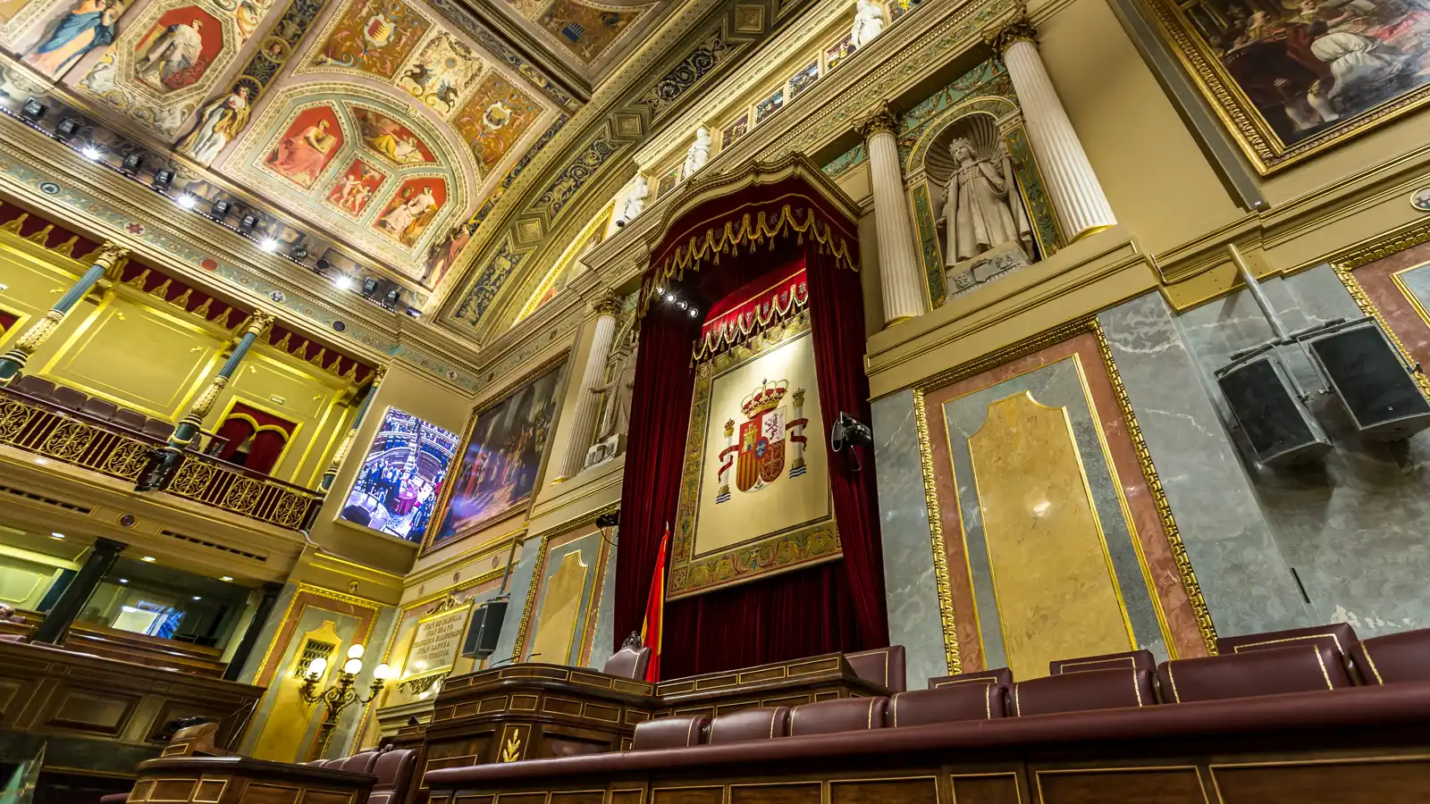 Interior del salón de plenos del Congreso de los Diputados fotografiado a contrapicado: el escudo de España bordado preside la tribuna bajo un dosel de cortinaje granate, con el techo de frescos dorados a la izquierda, columnas y estatuas a la derecha y las primeras filas de escaños de cuero granate en primer plano.