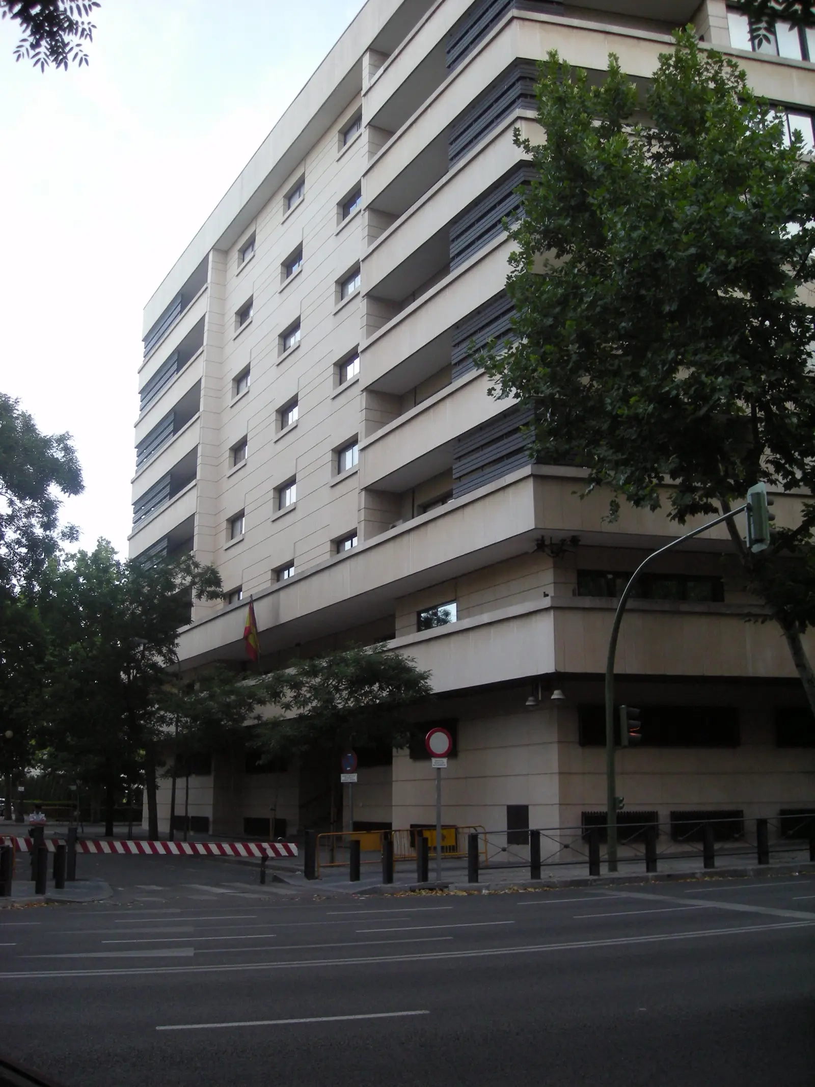 Edificio de siete plantas de piedra clara y celosías oscuras, fotografiado en esquina y a contrapicado desde la calzada; bajo el voladizo de la entrada ondea una bandera de España, y delante hay bolardos, vallas metálicas y árboles en la acera.