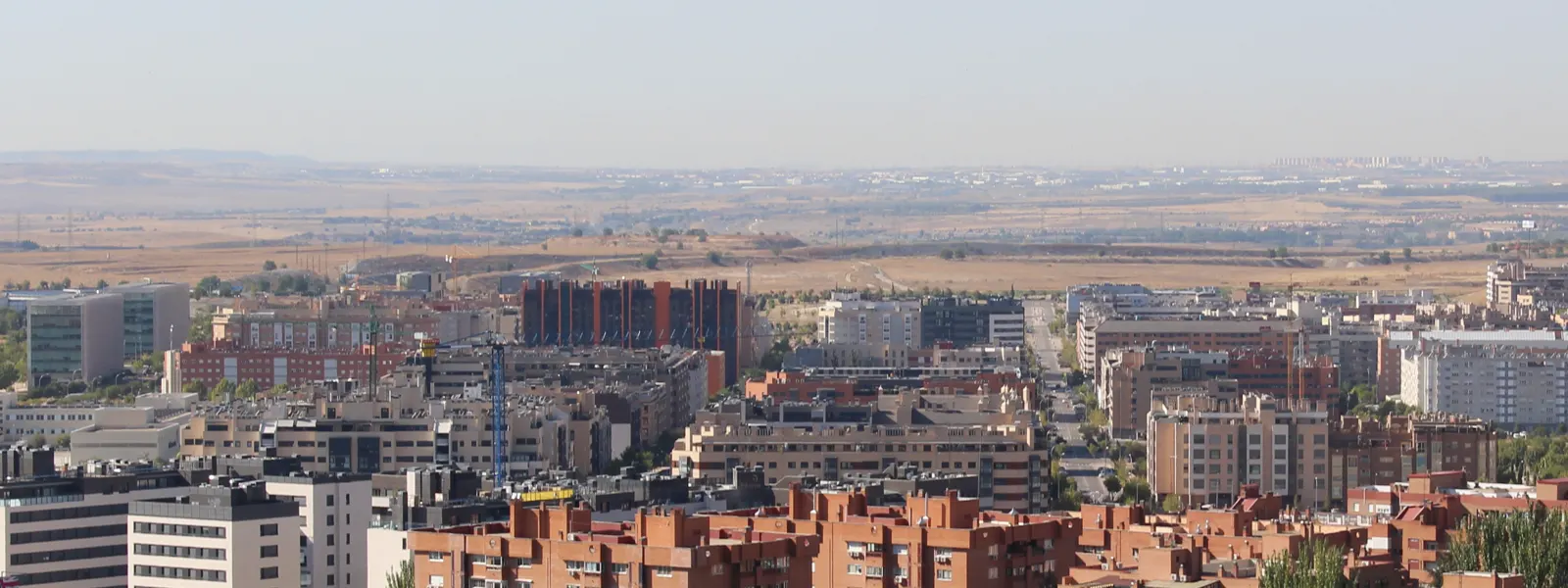 Vista panorámica de un barrio de bloques de viviendas de ladrillo rojo y fachadas claras, con dos grúas de obra entre los edificios y descampados sin urbanizar al fondo, bajo un cielo despejado.