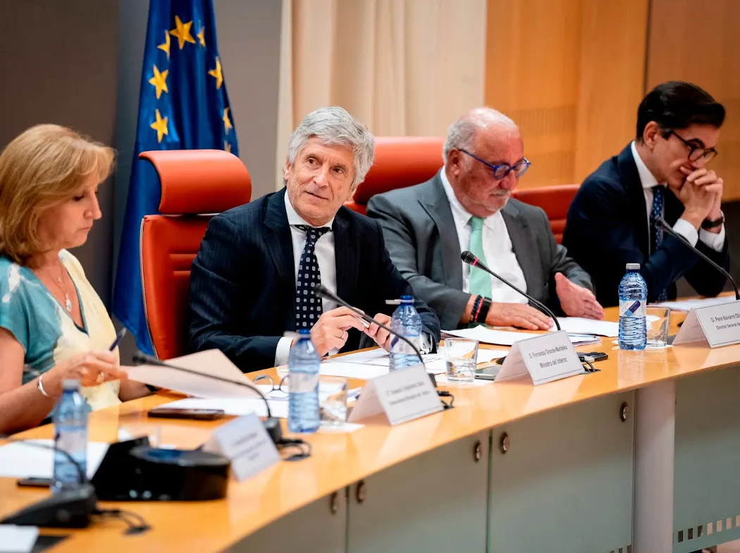 Cuatro personas sentadas tras una mesa curva de conferencia con micrófonos, carpetas y botellas de agua; delante de cada una hay un cartel identificativo y al fondo cuelga una bandera de la Unión Europea. Los carteles legibles corresponden al ministro del Interior, Fernando Grande-Marlaska, y al director general de Tráfico, Pere Navarro.