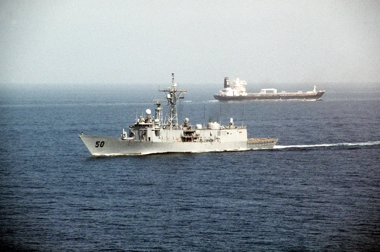 Una fragata de guerra gris con el número 50 en la proa navega dejando estela por un mar en calma; al fondo, entre la bruma, un petrolero cargado avanza en sentido contrario.