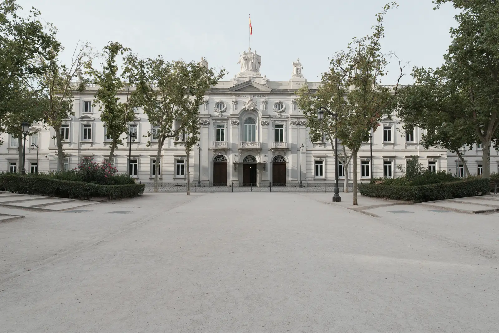 Fachada del Tribunal Supremo de España vista de frente desde la explanada de la plaza de la Villa de París: un edificio neoclásico de piedra clara con un frontón coronado por esculturas, la bandera de España ondeando sobre la cubierta, verjas negras al pie y plátanos de sombra a ambos lados.