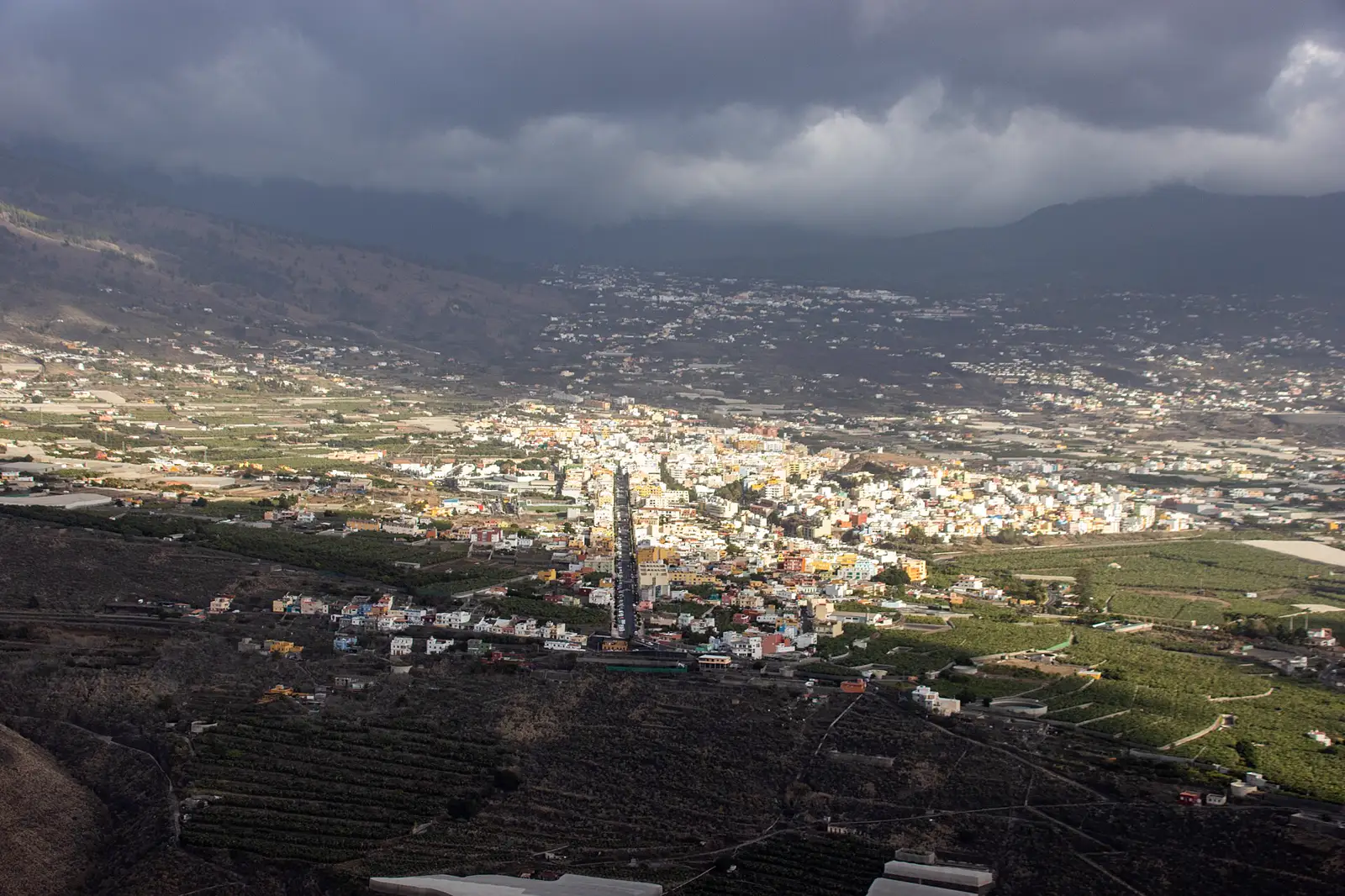 Vista en picado del valle de Aridane, en La Palma: el casco urbano de Los Llanos de Aridane, con casas blancas y de colores, rodeado de plataneras en terrazas y de terreno volcánico oscuro, bajo un cielo cubierto de nubes negras.