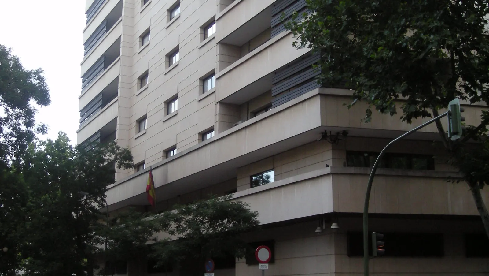 Esquina de un edificio de oficinas de piedra clara y siete plantas, con balcones corridos y celosías oscuras, visto desde la acera de enfrente entre árboles; bajo el voladizo de la entrada ondea una bandera de España.