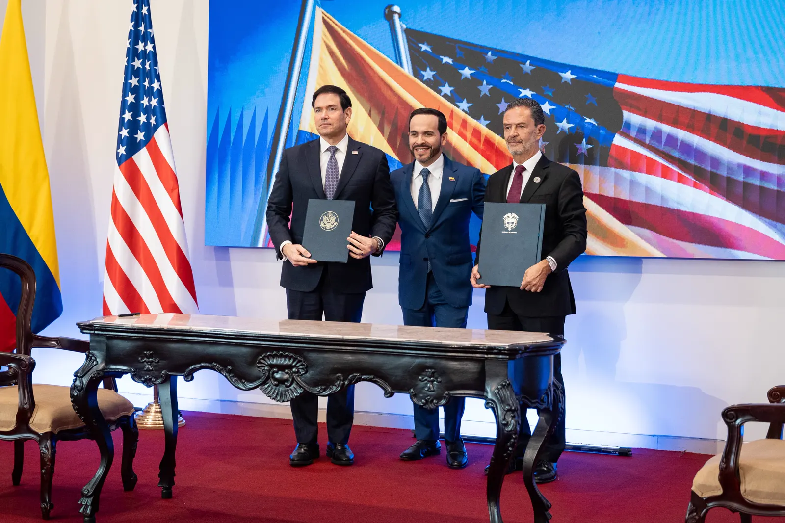 Tres hombres con traje posan de pie tras una mesa de firma sosteniendo dos carpetas, una con el sello de Estados Unidos y otra con el de Colombia, ante las banderas de ambos países.