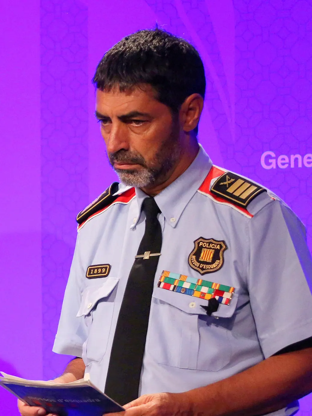 Josep Lluís Trapero, con el uniforme celeste de los Mossos d'Esquadra y galones de mayor, mira unos papeles ante un fondo violeta con el logotipo de la Generalitat.